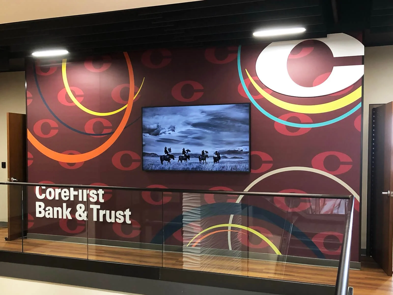 Interior wall of a bank with colorful abstract circular patterns, a TV displaying a black and white photo of horseback riders, and a logo that reads 'CoreFirst Bank & Trust' in white text.