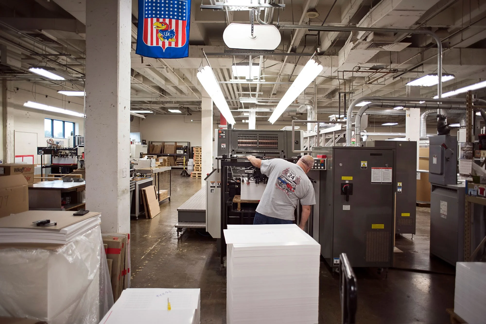 A worker operating a printing or manufacturing machine in a large industrial workspace with shelves of supplies and other equipment in the background.