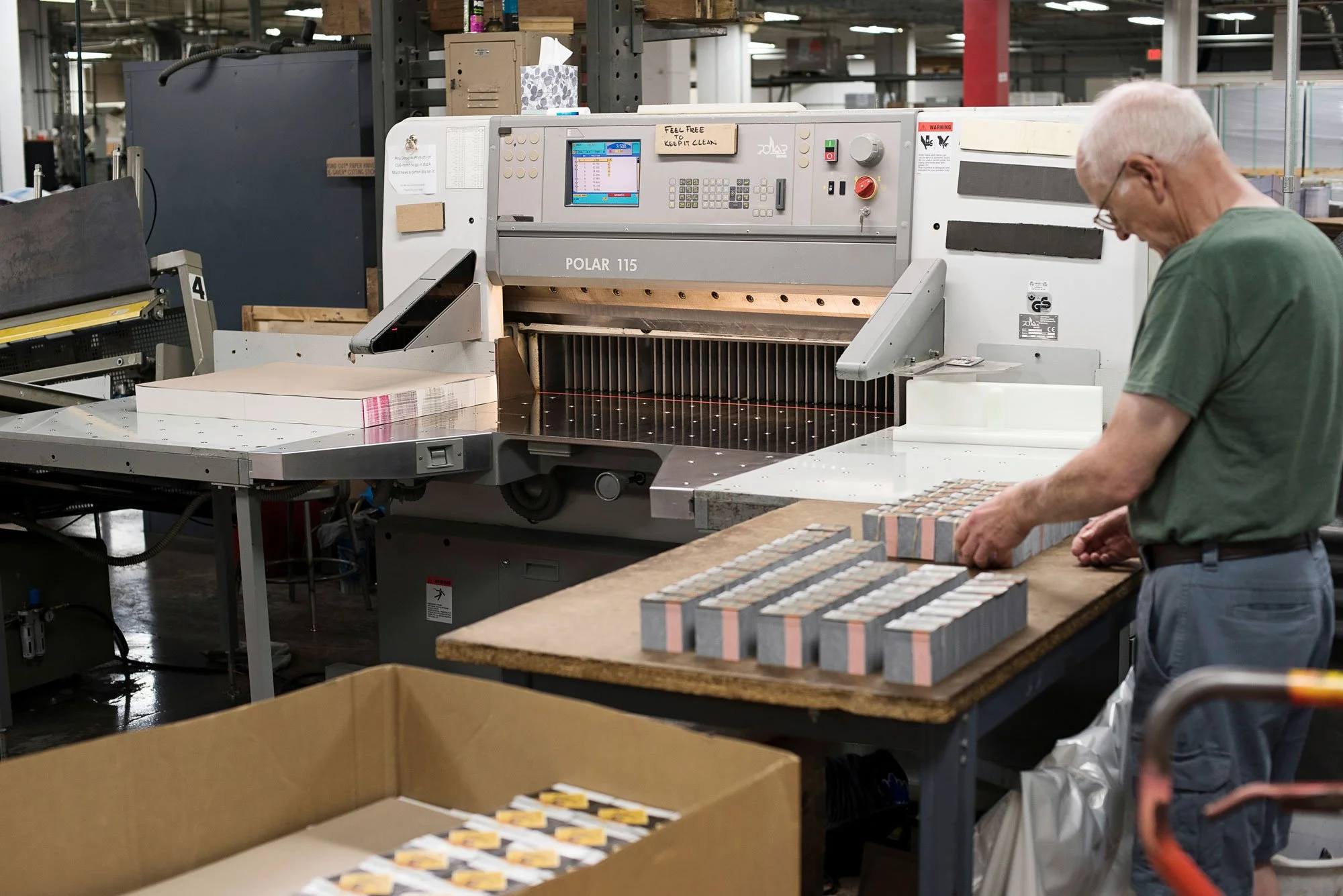 An older man with glasses inspects ceramic tiles on a workbench inside a tile manufacturing factory, with a large industrial cutting machine labeled 'POLAR 115' in the background.