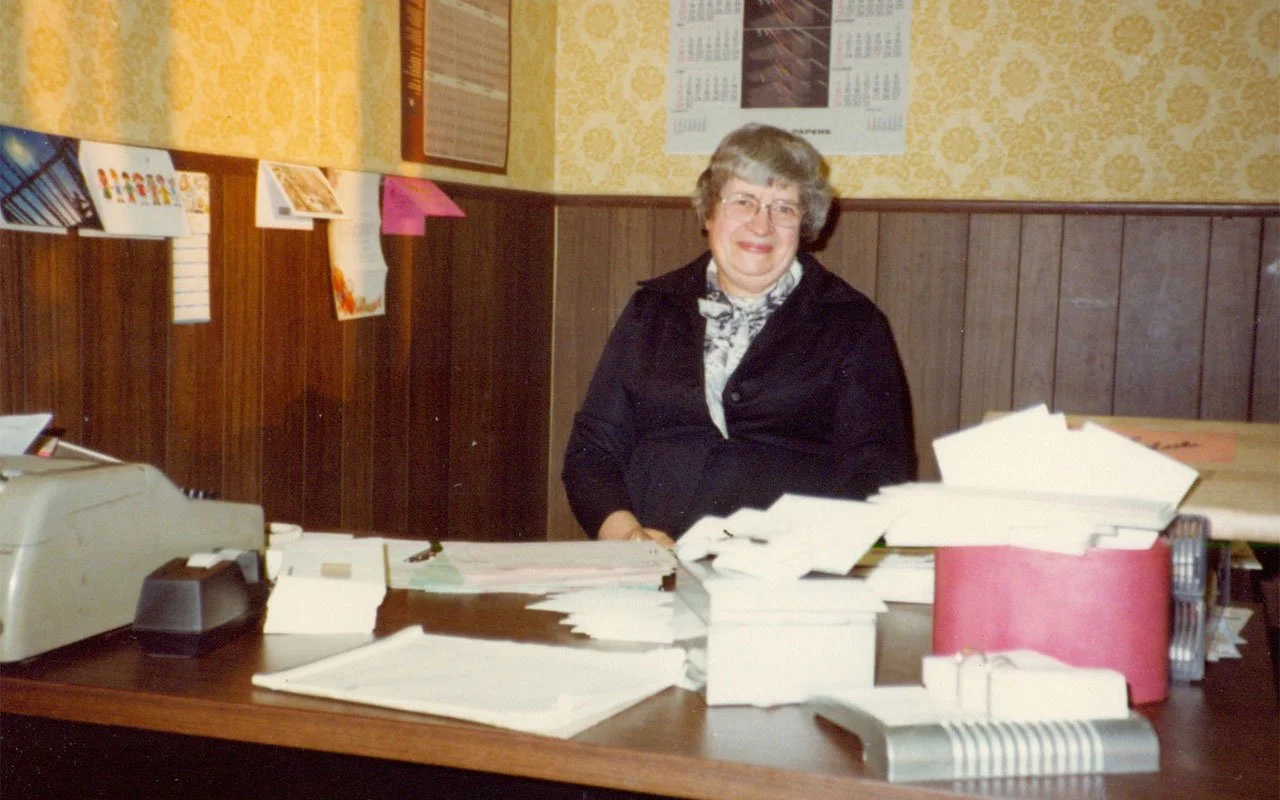 A smiling woman standing behind a cluttered desk with papers and office supplies in a room with wood-paneled walls and yellow patterned wallpaper.