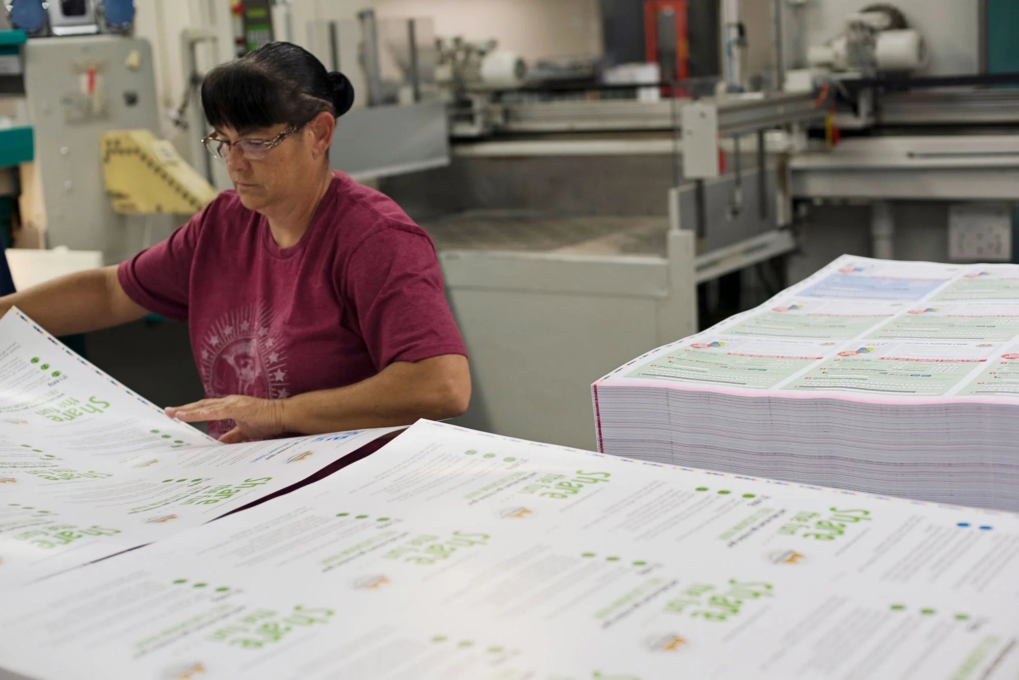 A woman with dark hair, glasses, and a maroon t-shirt sitting at a work table with large stacks of printed materials, possibly flyers or brochures, in what appears to be a printing or assembly facility.