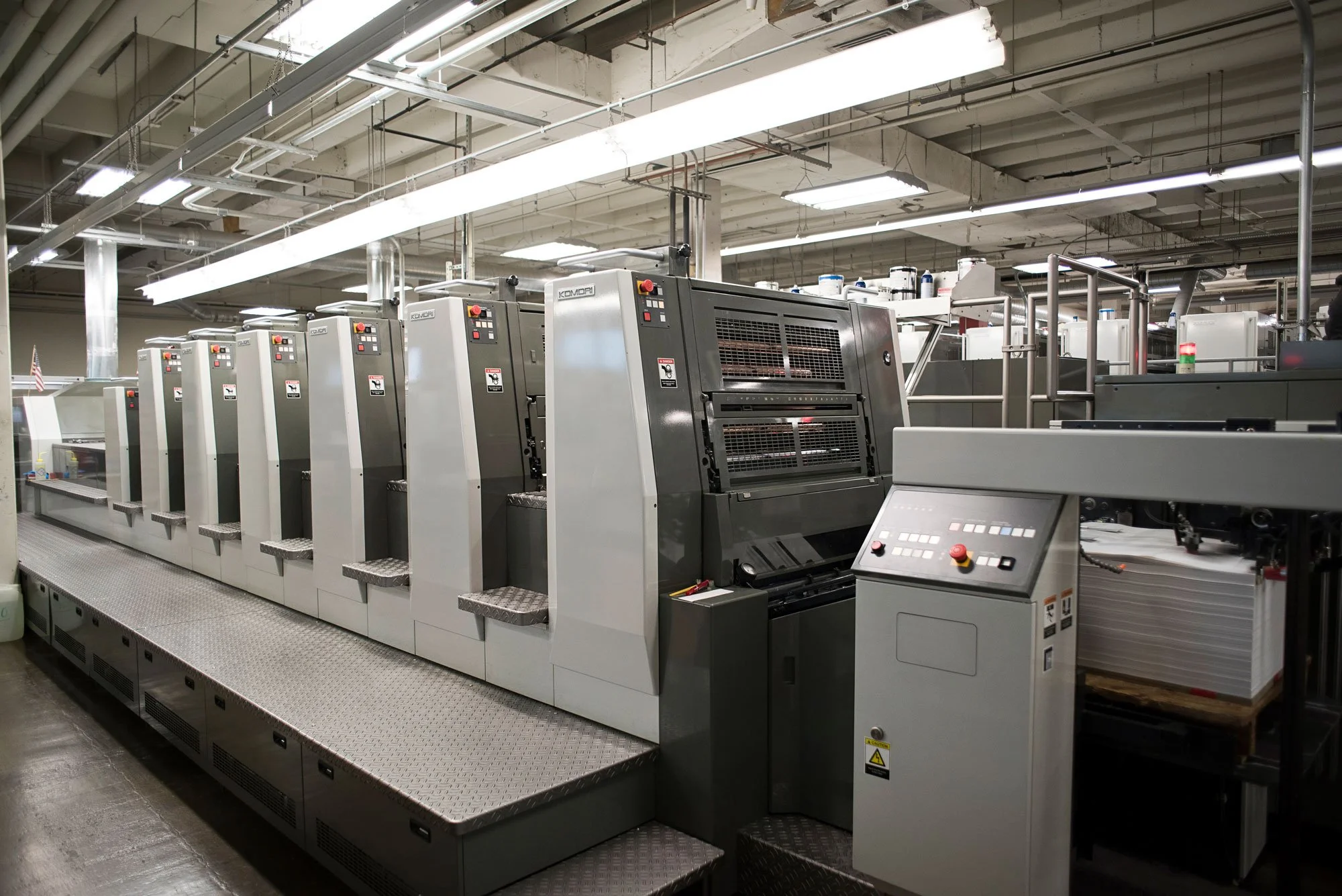 Multiple industrial printing presses in a factory, with control panels and paper stacks, under bright fluorescent lighting.