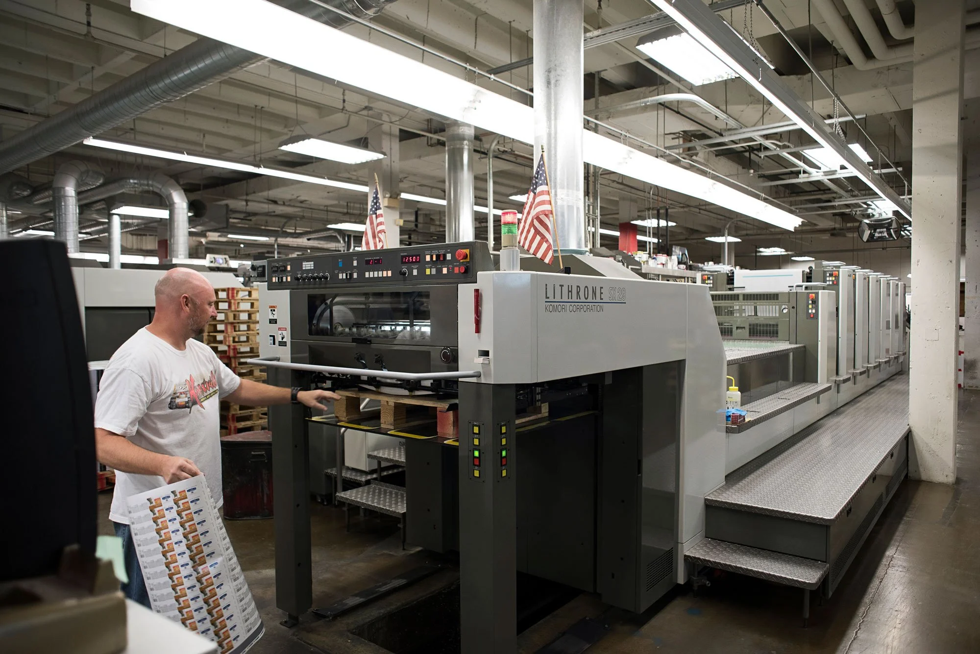 A worker in a T-shirt operating a large industrial printing machine in a factory setting, with American flags displayed above and rows of equipment in the background.