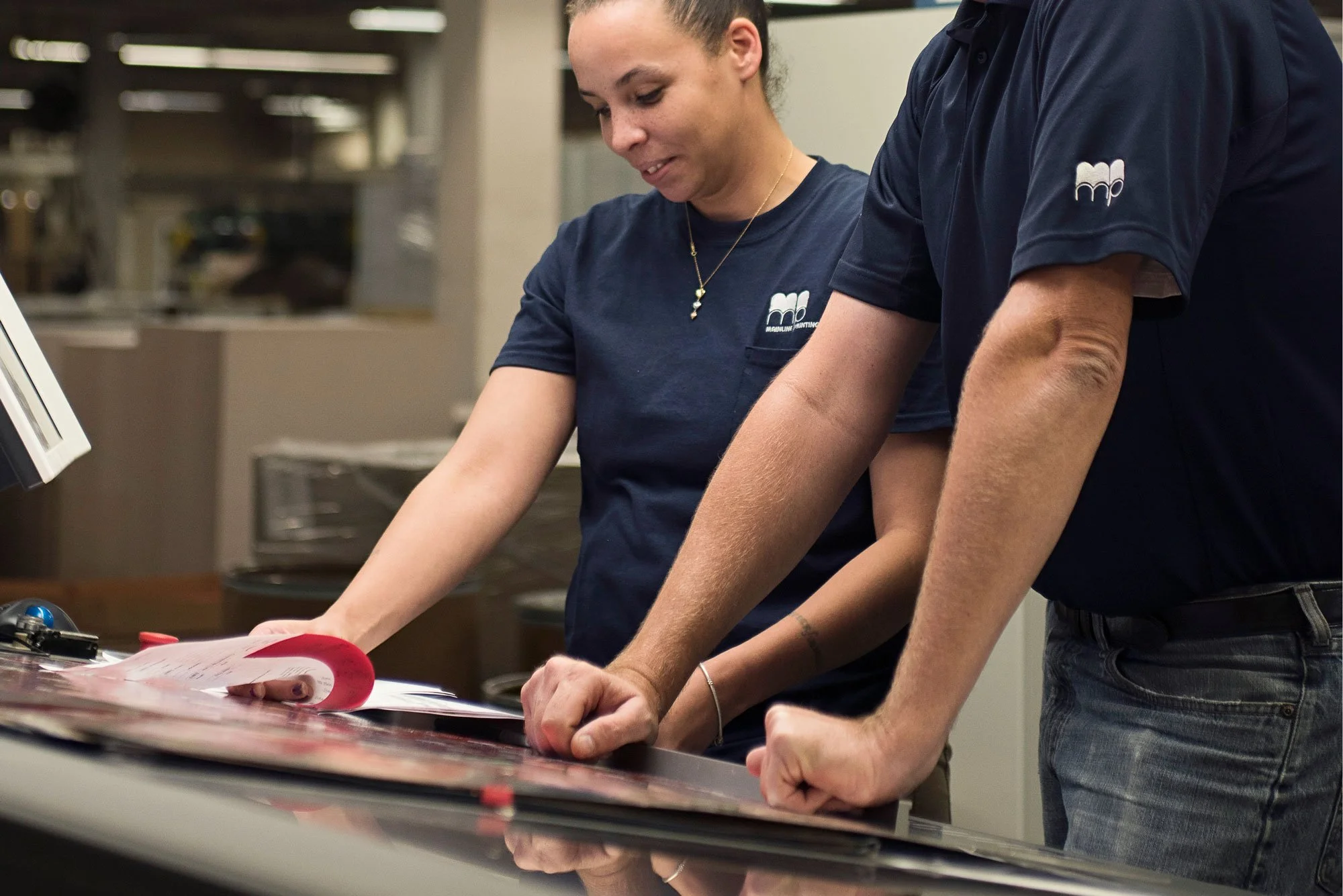 Two people wearing navy blue shirts working with documents on a counter in a commercial print shop.