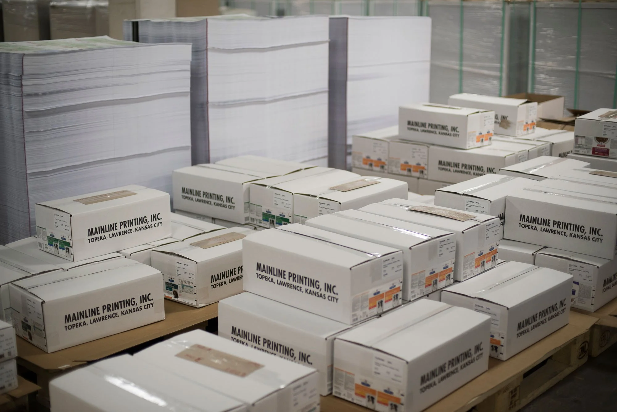 Boxes labeled 'Mainline Printing, Inc., Topeka, Lawrence, Kansas City' stacked on wooden pallets in a printing warehouse.
