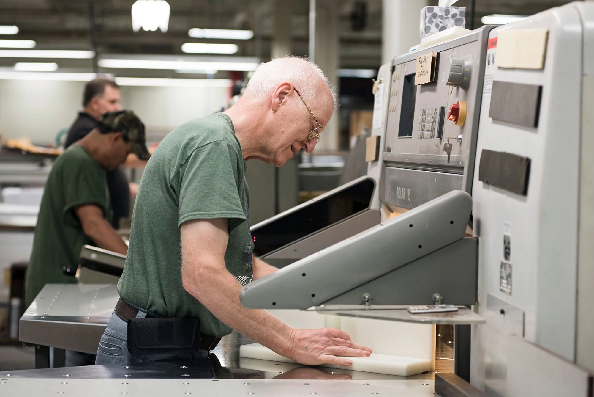 A man wearing a green shirt and glasses operating a commercial machine in a factory or workshop environment, with other workers in the background.