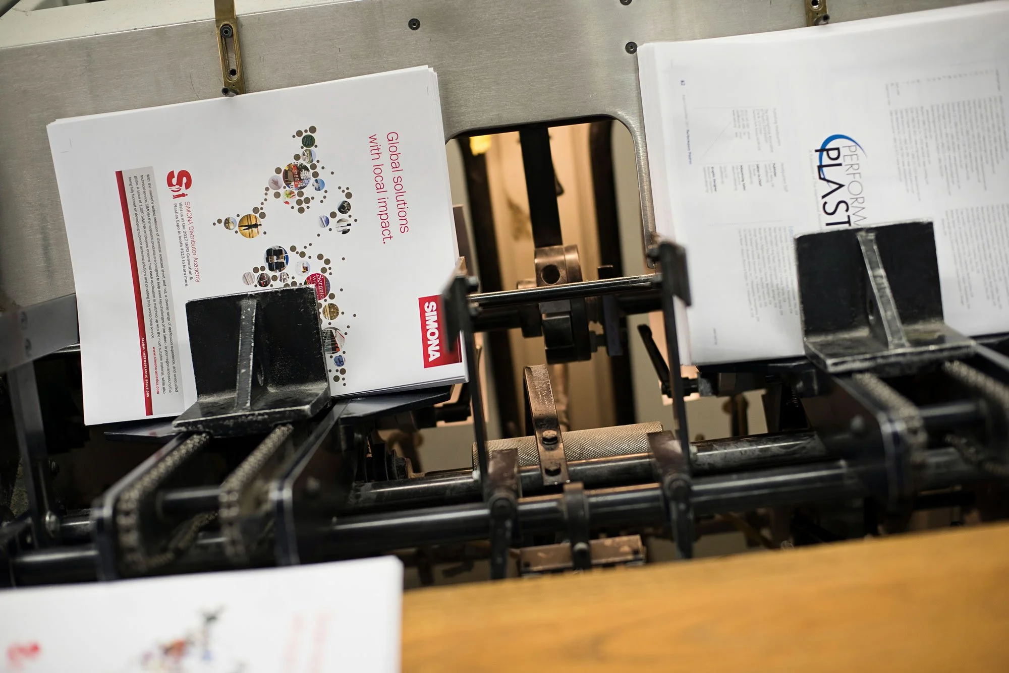 A printing machine producing brochures, with two brochures visible, one promoting shown as 'Swanson' with a slogan 'Global solutions with local impact' and another with the title 'PERFORMANCE PLUS'.