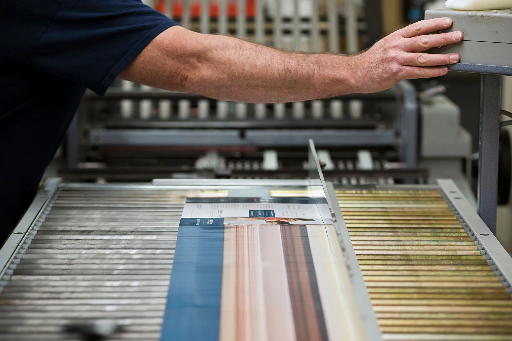 A person's arm and hand adjusting a printing press with fabric samples underneath, in a textile or print manufacturing setting.