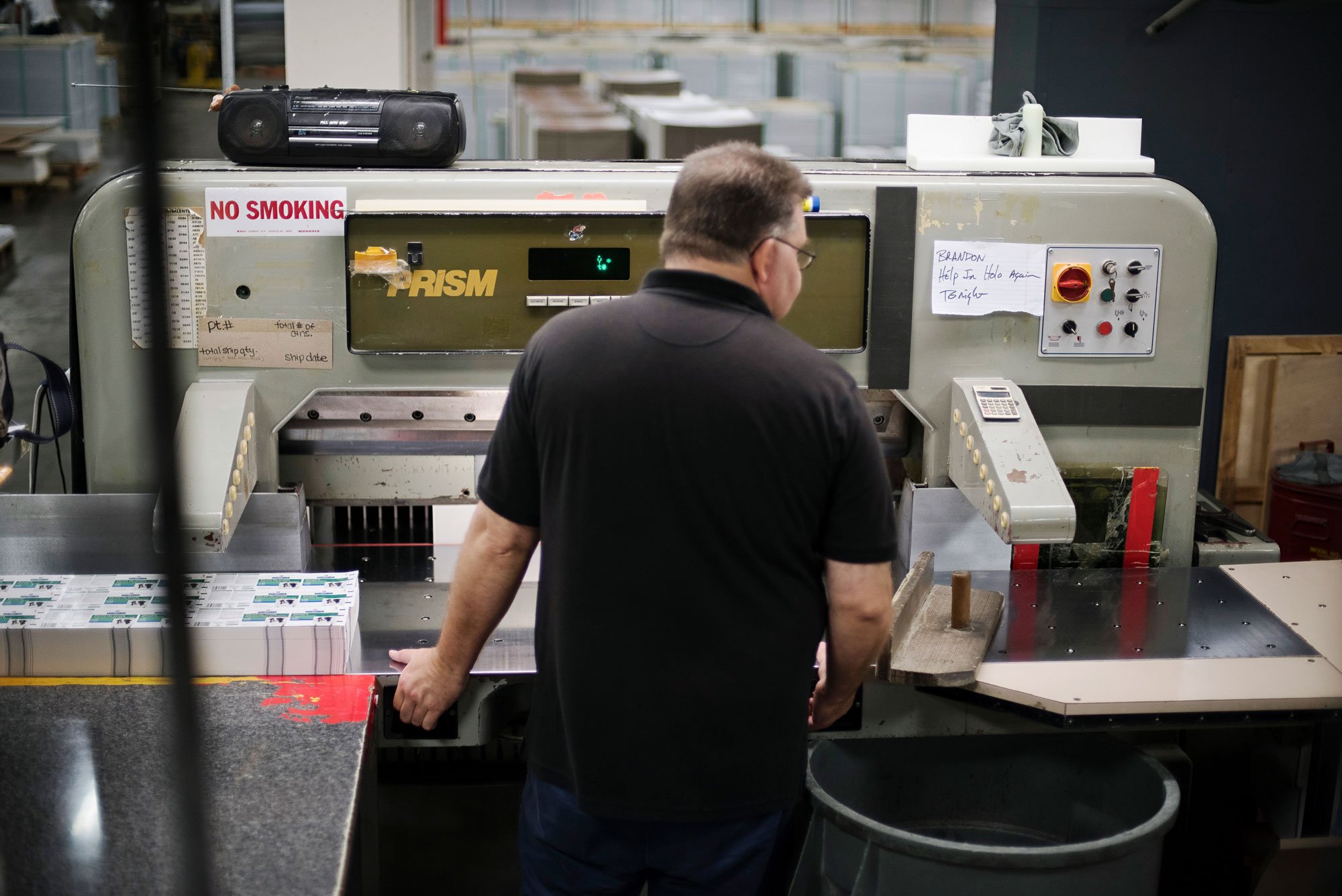 A man in a black shirt working in a factory or warehouse, standing in front of a large industrial machine.