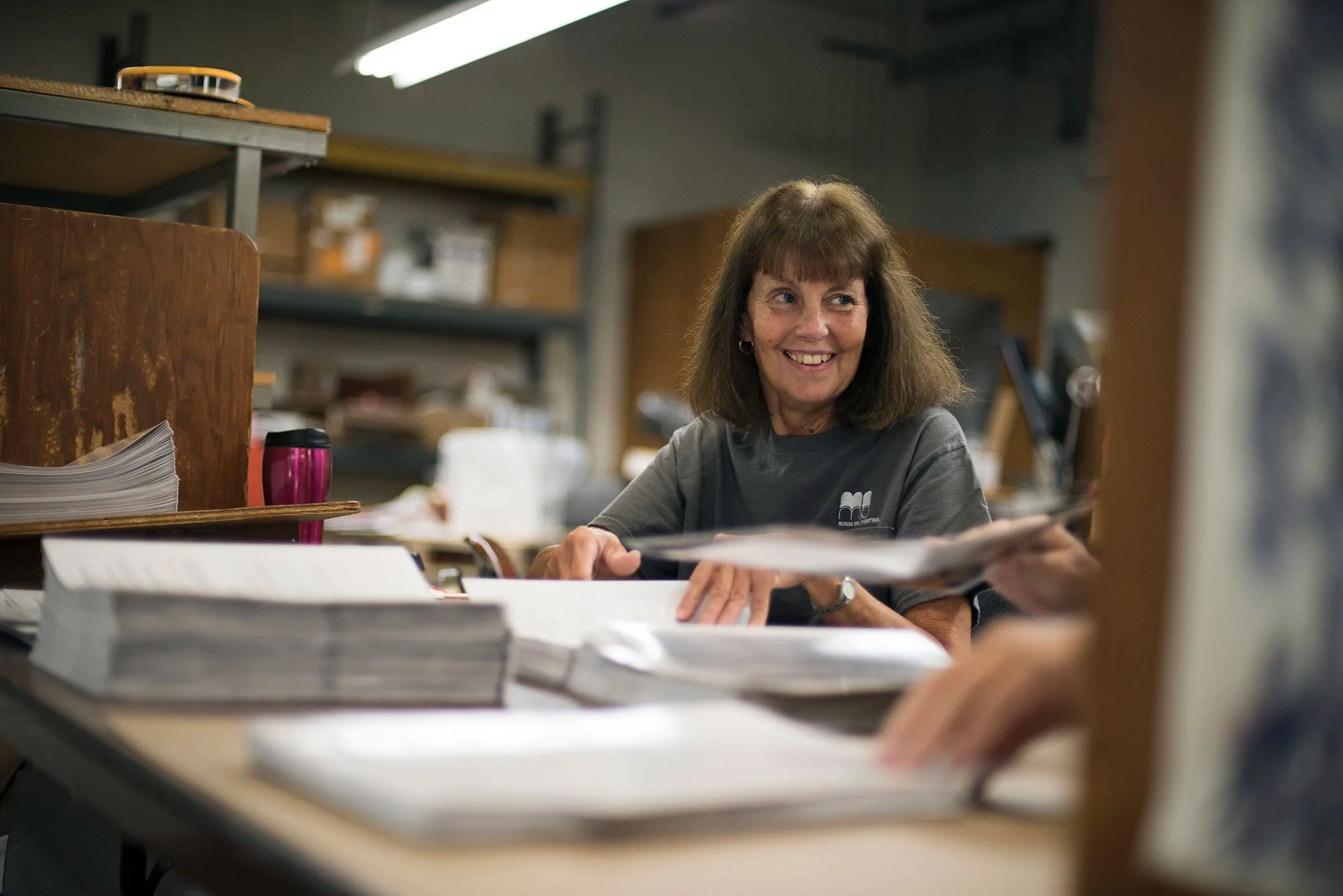A woman smiling and engaging in conversation while sitting at a desk filled with stacks of papers and documents in a workshop or office setting.