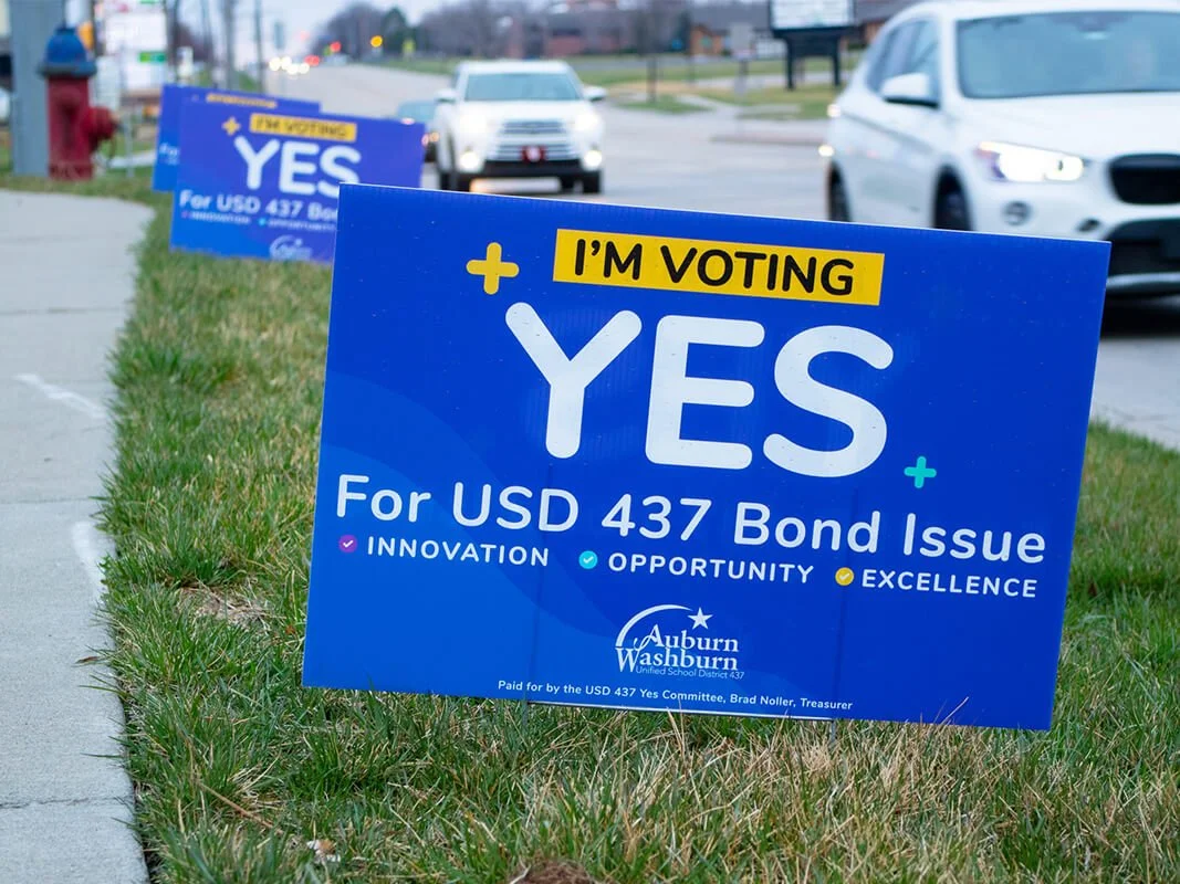 Political campaign signs on grass along sidewalk, advocating voting yes for USD 437 bond issue, with cars on street in background.