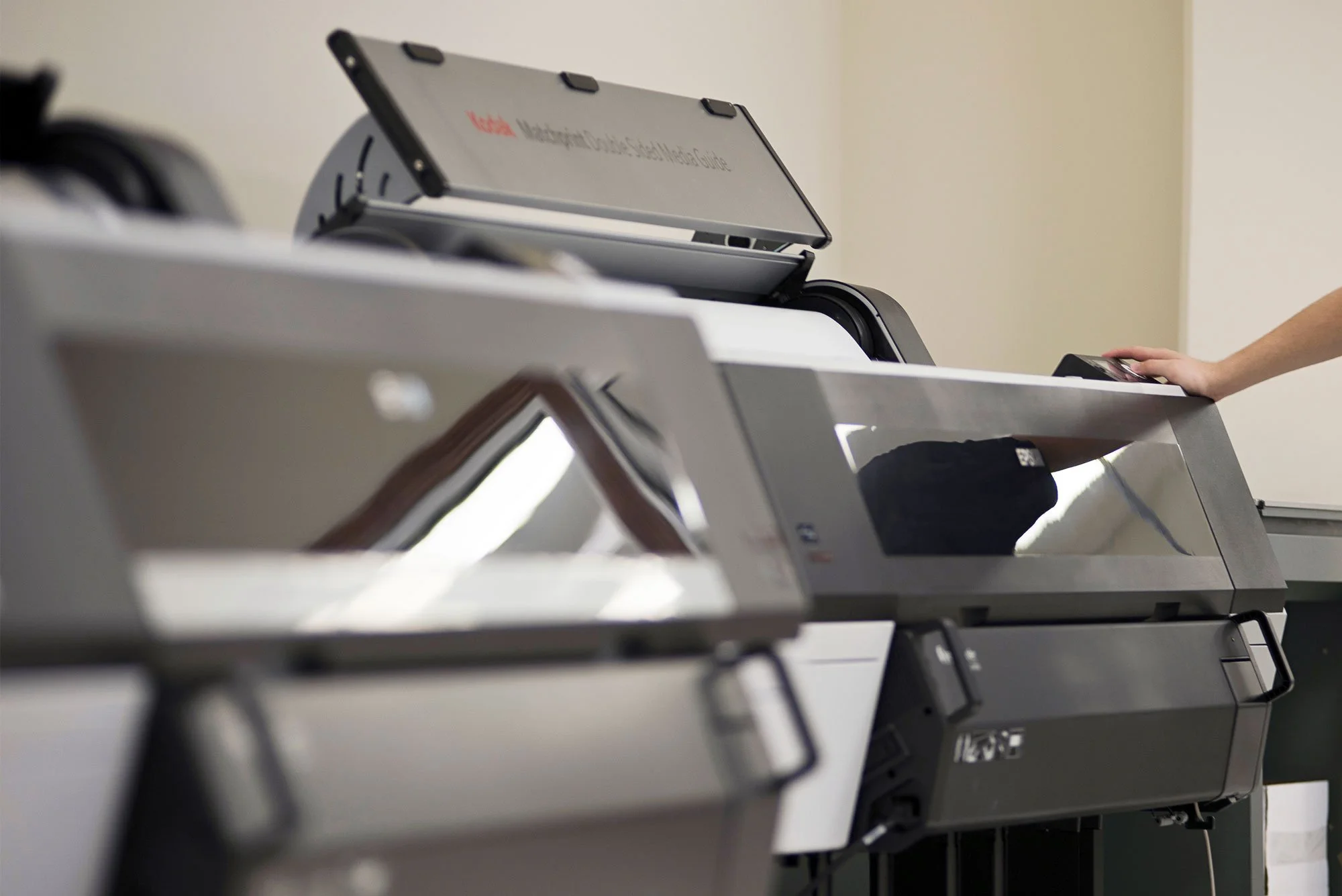 A person on the right is operating a large photocopier or scanner in a room with plain walls, with multiple pieces of office equipment visible.