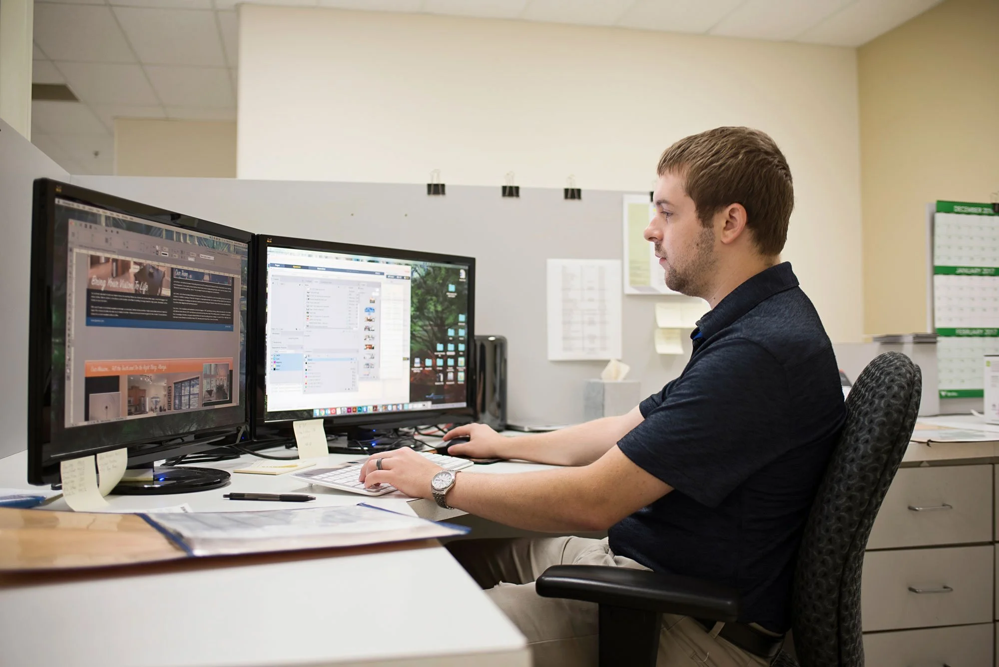 Man working at dual computer monitors on office desk, with papers, pens, and sticky notes.
