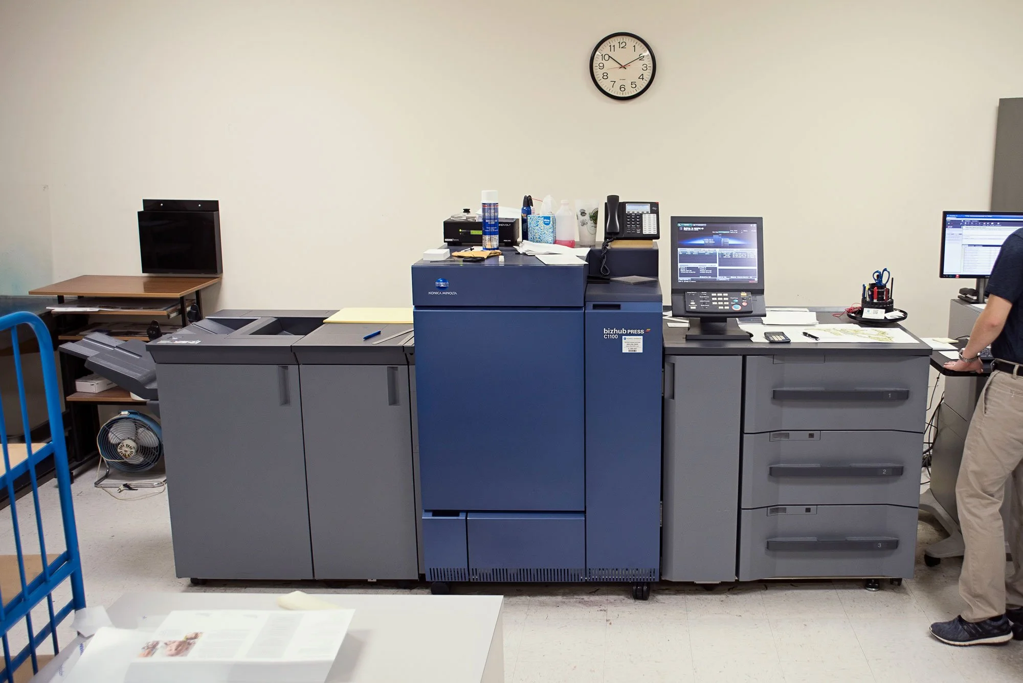 Office photocopier and printer machine with desktop monitors, clocks, and office supplies on top, in a corporate office setting.