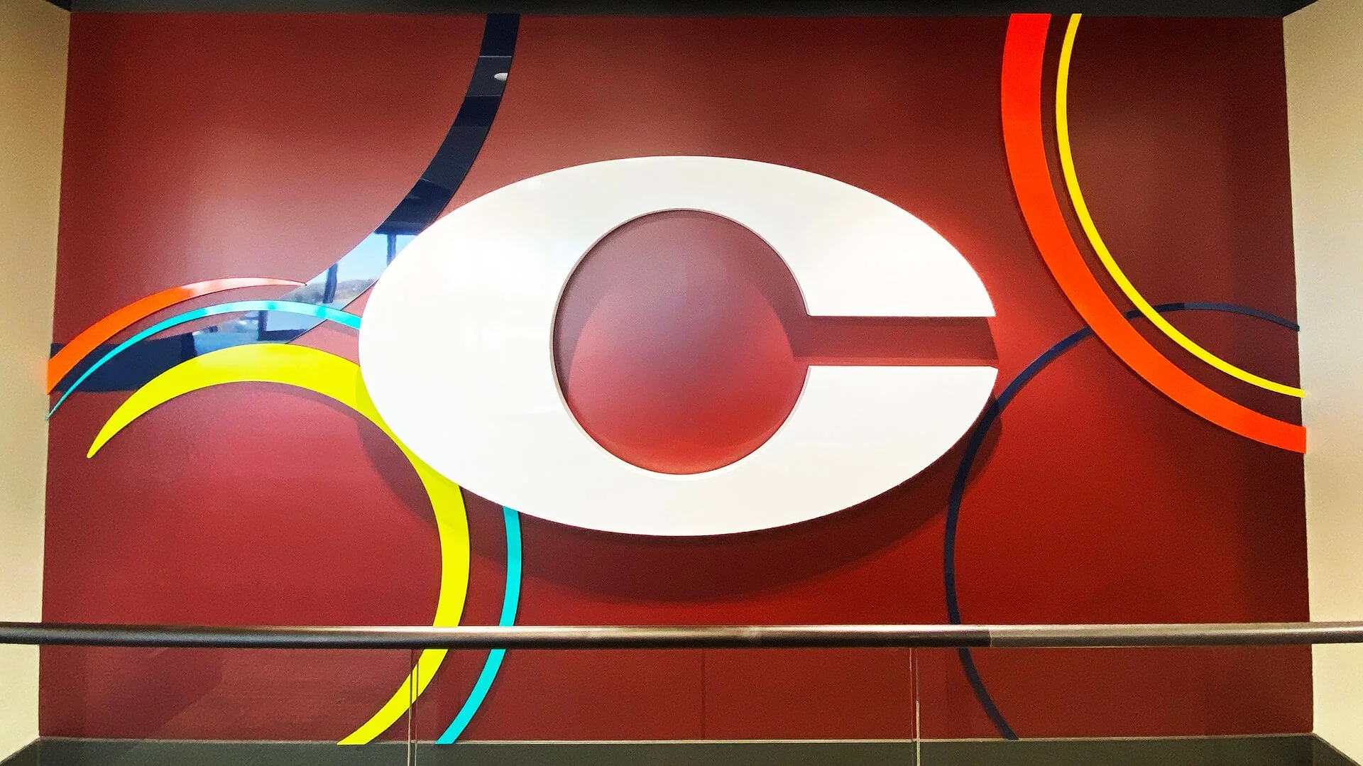 A large abstract wall art piece featuring a white oval shape with a cut-out center on a red background with curving, multicolored lines in yellow, blue, orange, and black. The piece appears to be three-dimensional and is mounted on the wall of a building interior.
