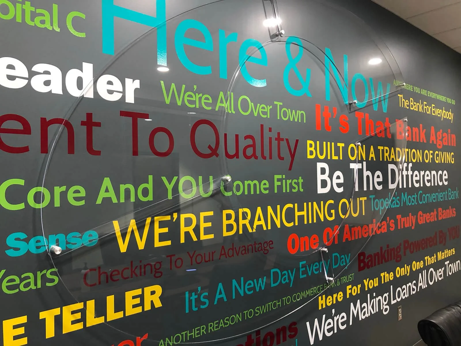 Colorful wall display with various banking slogans and phrases in different font sizes and colors, including words like 'Here & Now,' 'We’re Branching Out,' 'Core And YOU Come First,' and 'One Of America’s Truly Great Banks.'