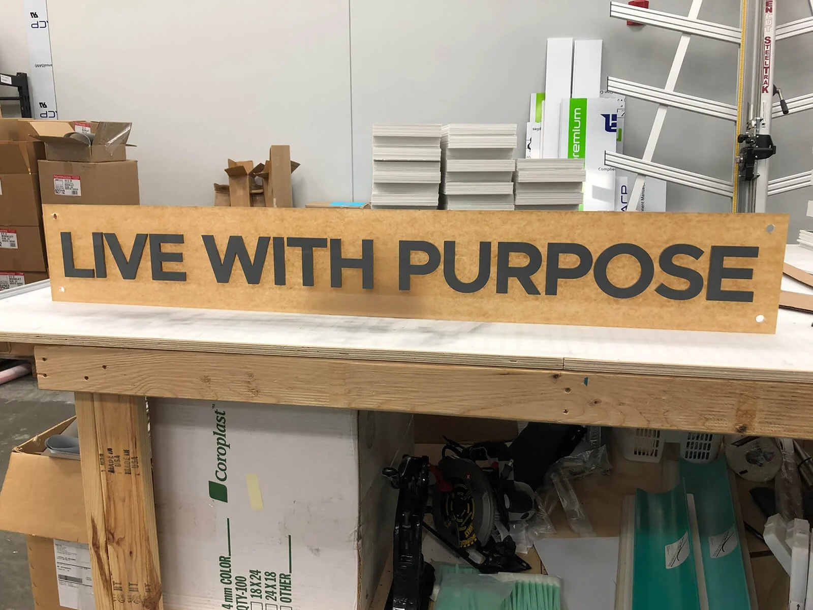A wooden sign with black text that says 'LIVE WITH PURPOSE' on a workbench in a storage or workshop area, with boxes and supplies in the background.