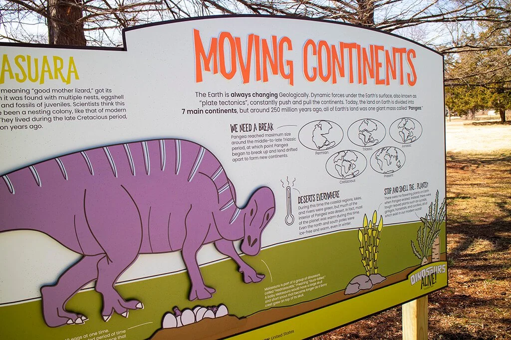Educational sign about moving continents featuring a cartoon purple dinosaur with eggs underneath, surrounded by desert plants and trees in the background.