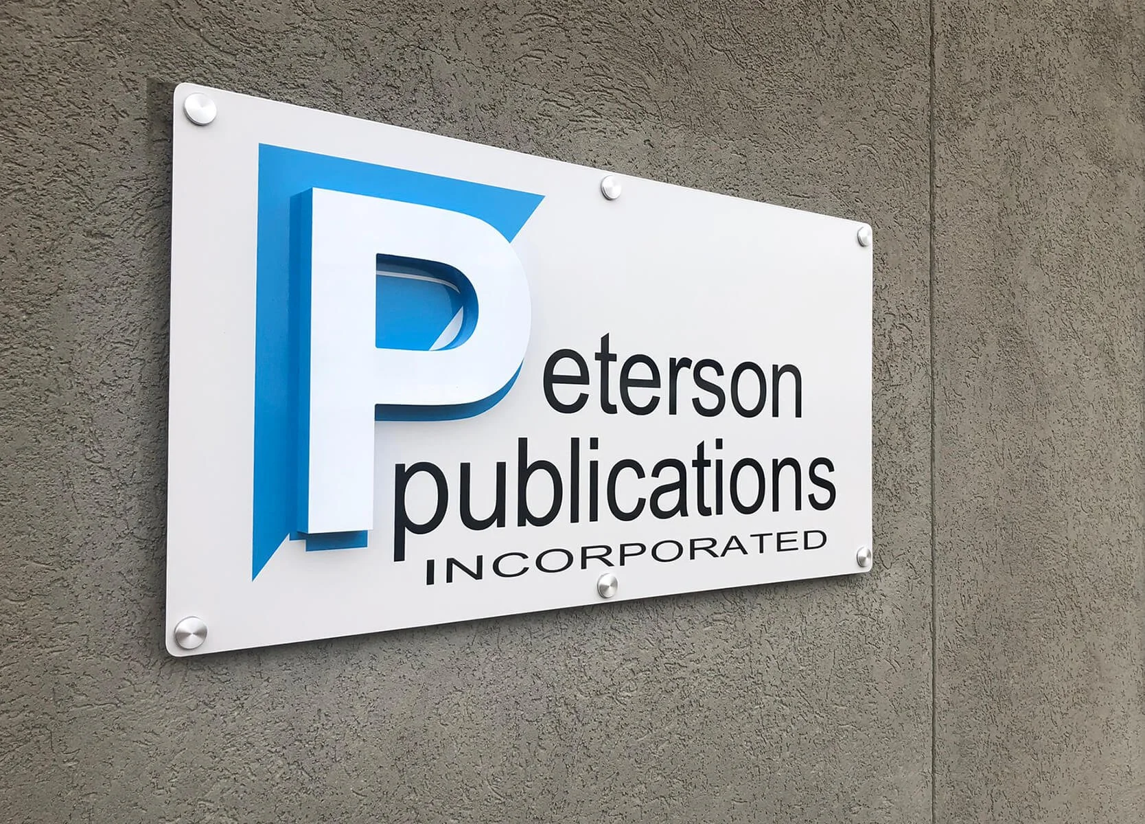 Sign for Peterson Publications Incorporated on a gray wall, featuring a large blue and white 3D letter 'P' and black text with the company name.