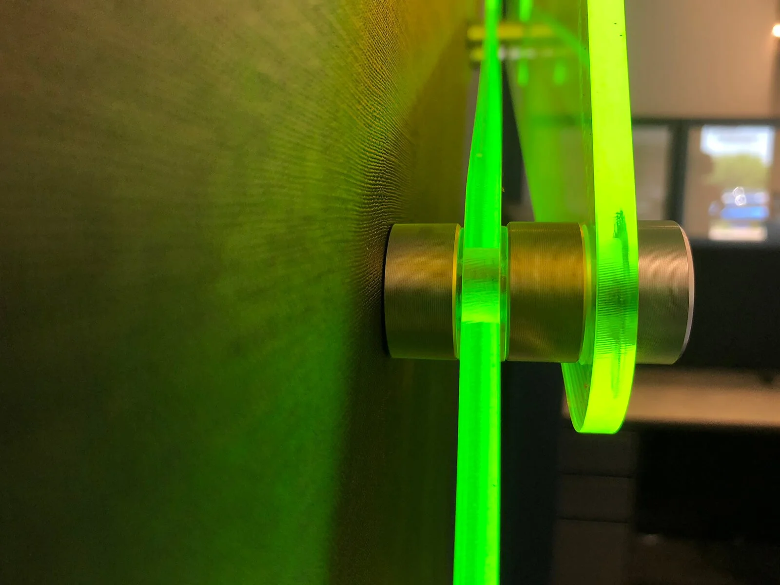 Close-up of a neon green LED strip light wrapped around a metallic cylindrical object on a wooden surface inside a room, with blurred background visible through a window.