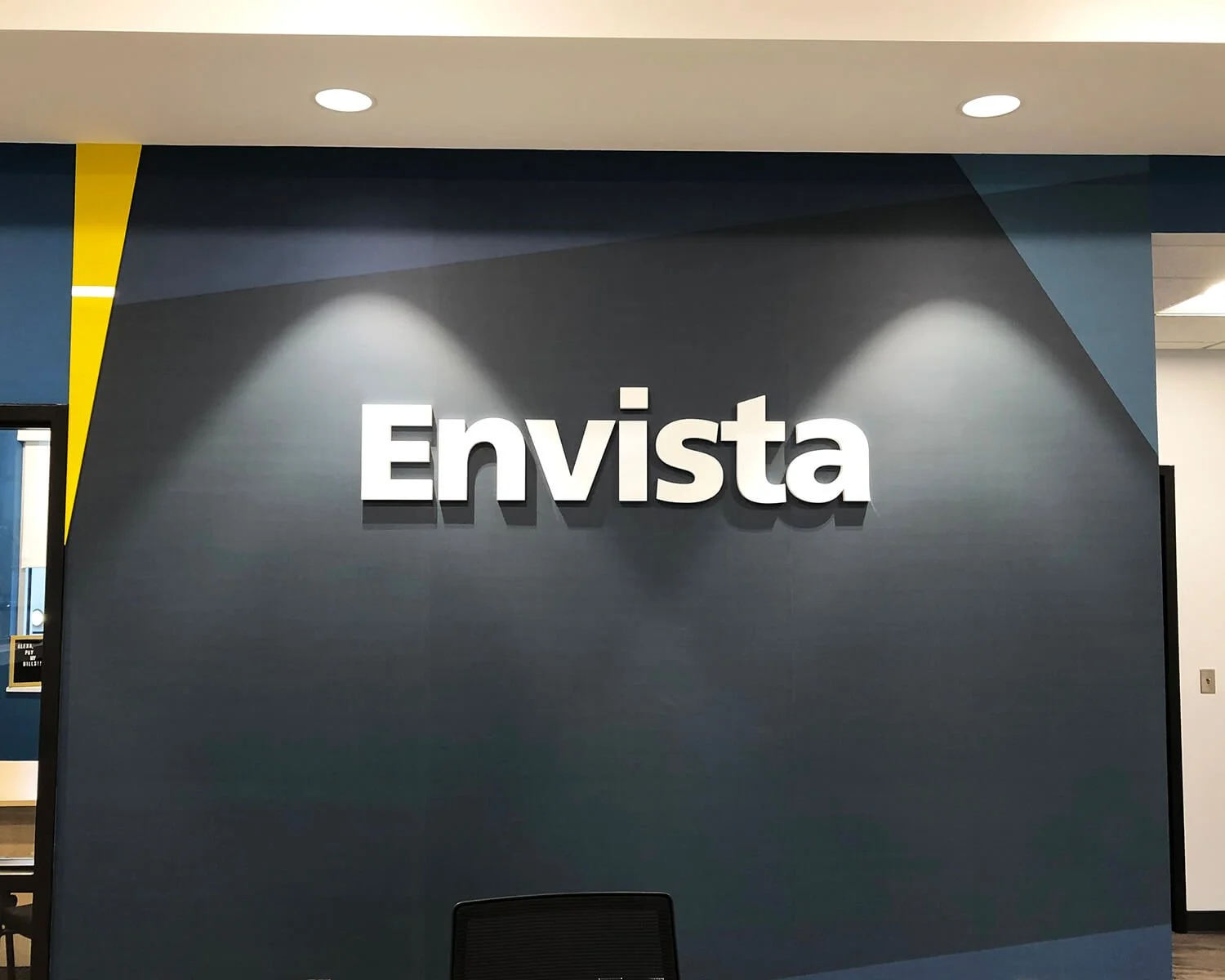 Interior office wall with the word 'Envista' in white 3D letters, illuminated by two spotlights on a dark gray wall.