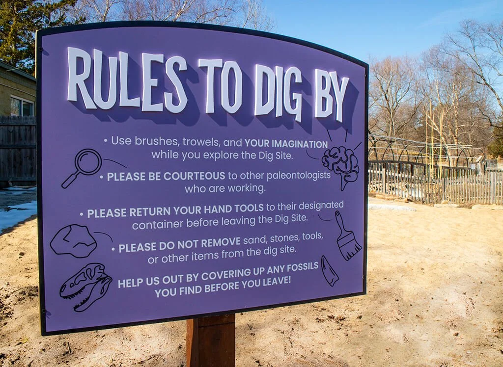 A sign at a dig site titled "Rules to Dig By" with rules for paleontology etiquette, including using brushes, being courteous, returning tools, not removing items, and helping cover fossils, with illustrations of a magnifying glass, brain, paintbrush