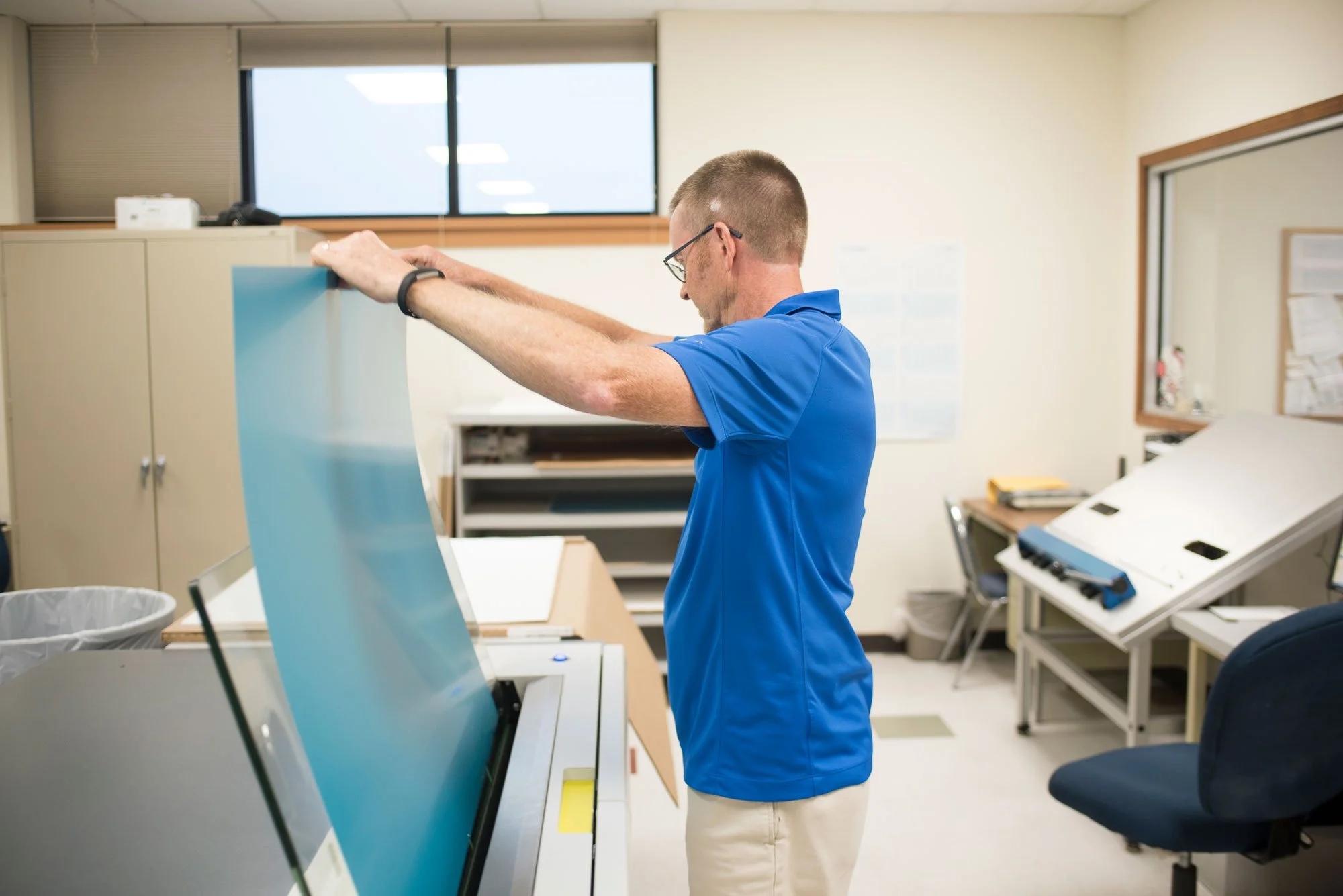 A man in a blue shirt and beige pants standing in an office or laboratory, opening a large equipment or machine with a clear blue cover.