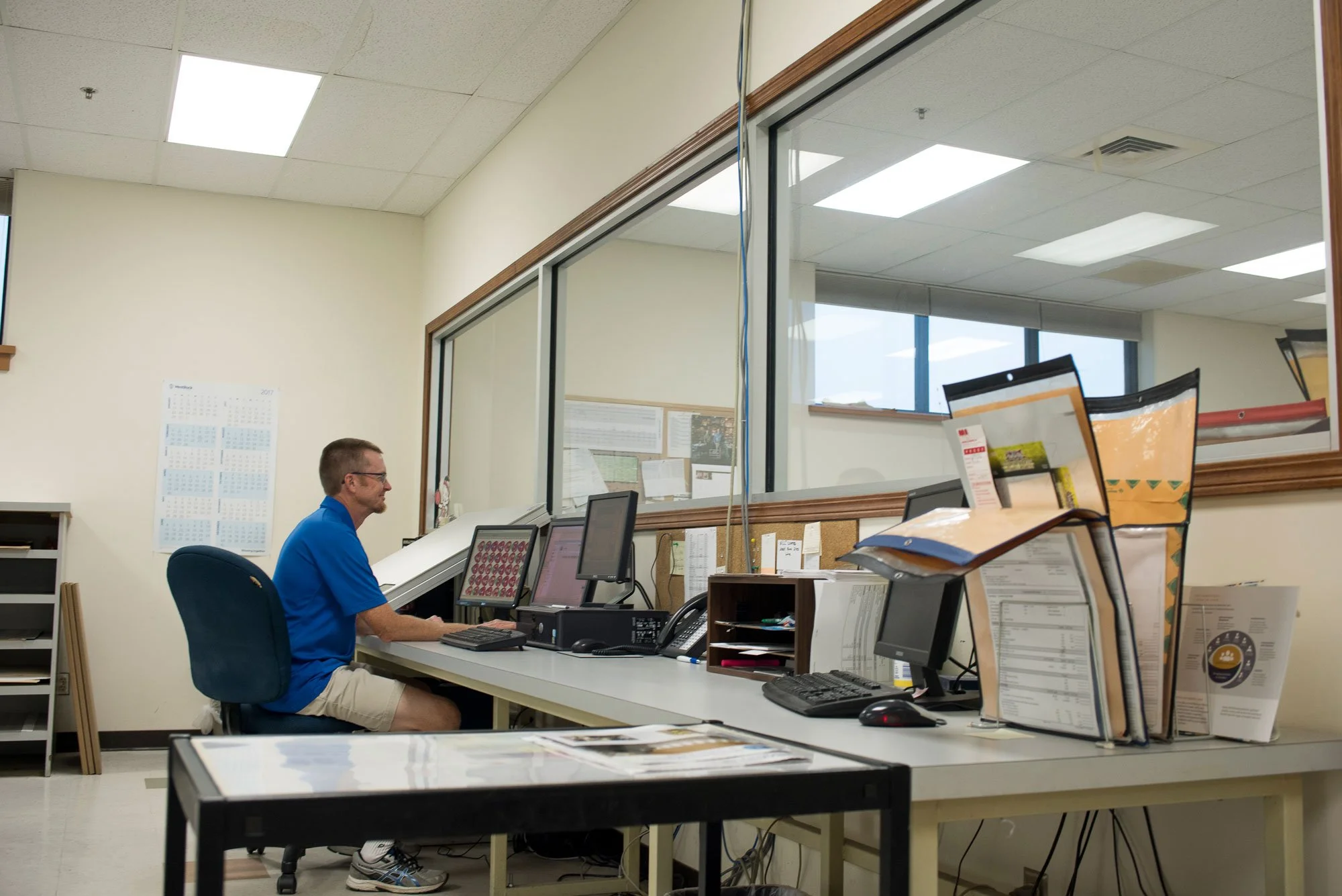 Man in a blue shirt and khaki shorts working at a computer desk in an office, with multiple monitors, papers, and office supplies on the desk.