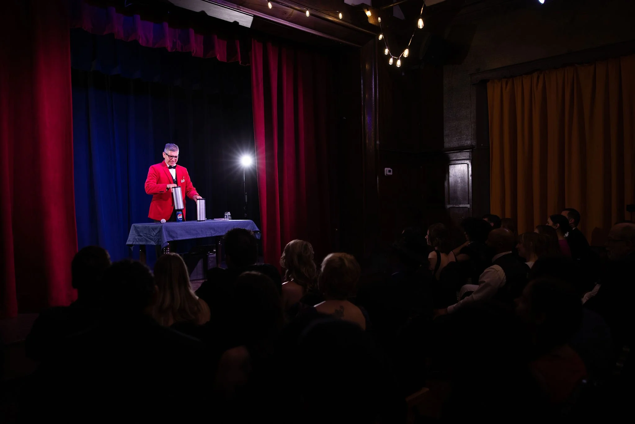 Cleveland magician in a red blazer with a bowtie performing a magic trick on a stage with a dark blue and red curtain background. The audience is watching from the darkened room.