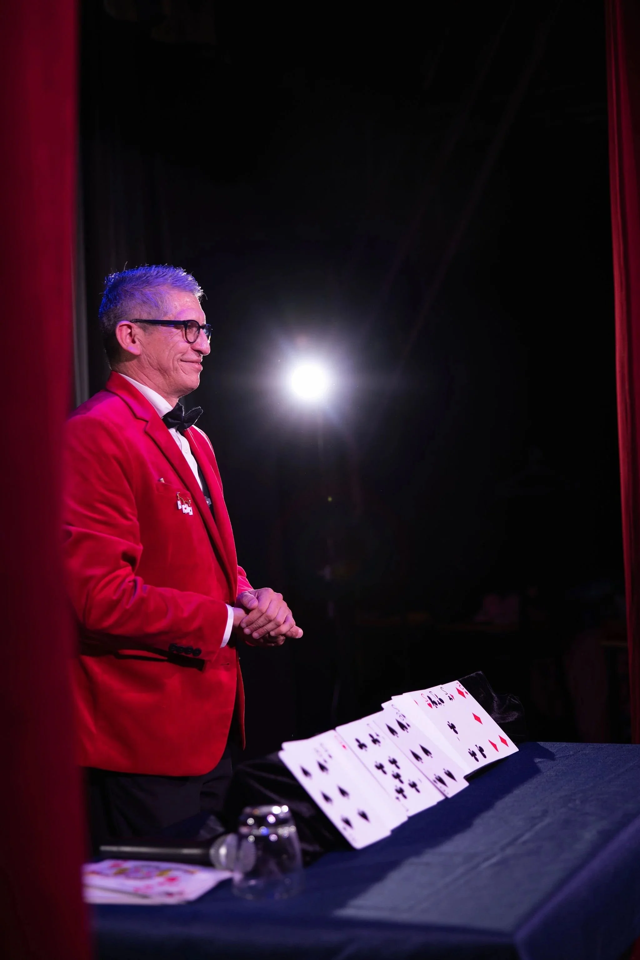 Cleveland magician David is standing with closed eyes and clasped hands on a stage. He is surrounded by red curtains, with a bright light source behind him. Playing cards are displayed on a table in front of him.