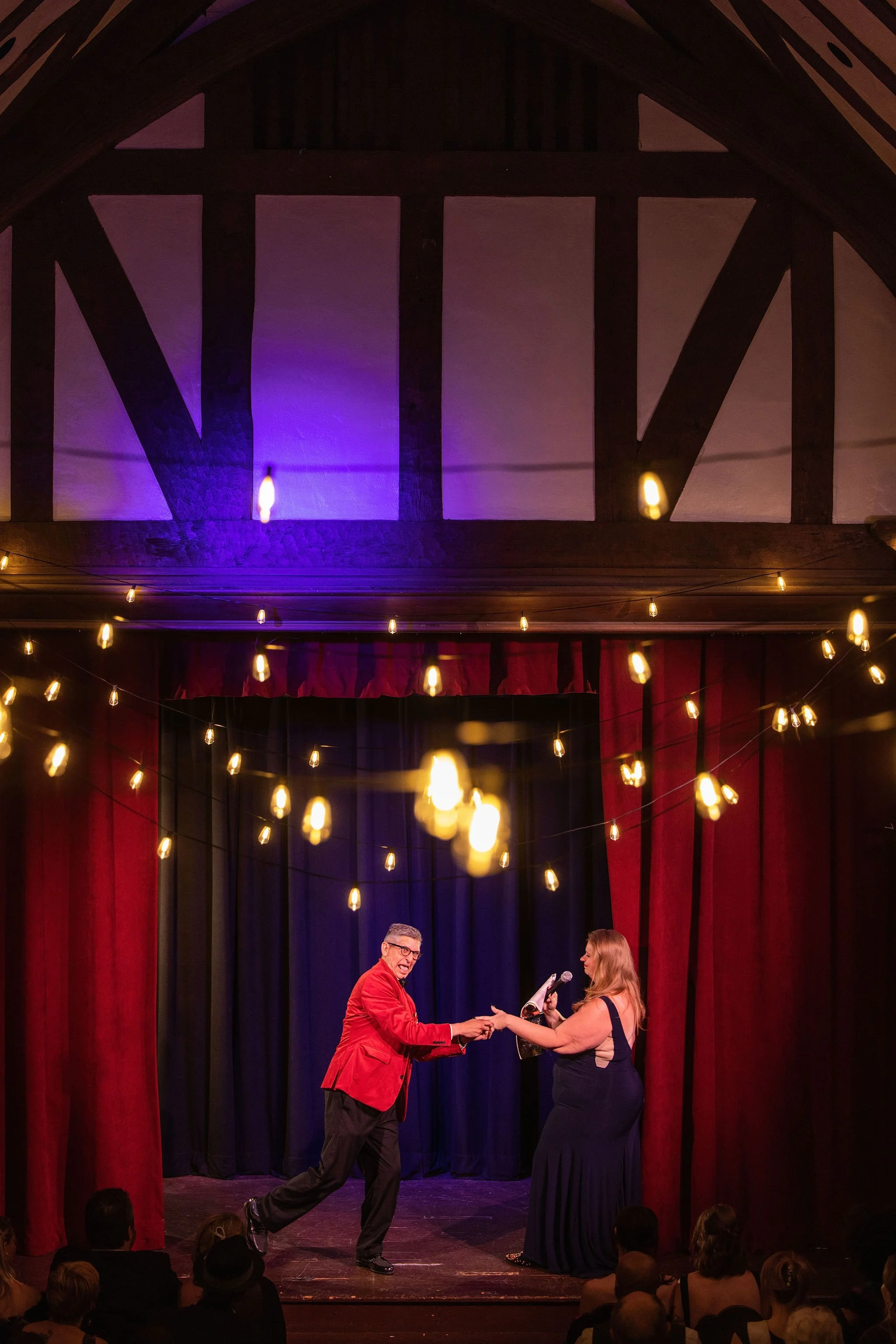 David Lee n a red suit hands a plaque or award to a woman in a black gown on a stage with red and blue curtains, string lights hanging above, and an audience watching.