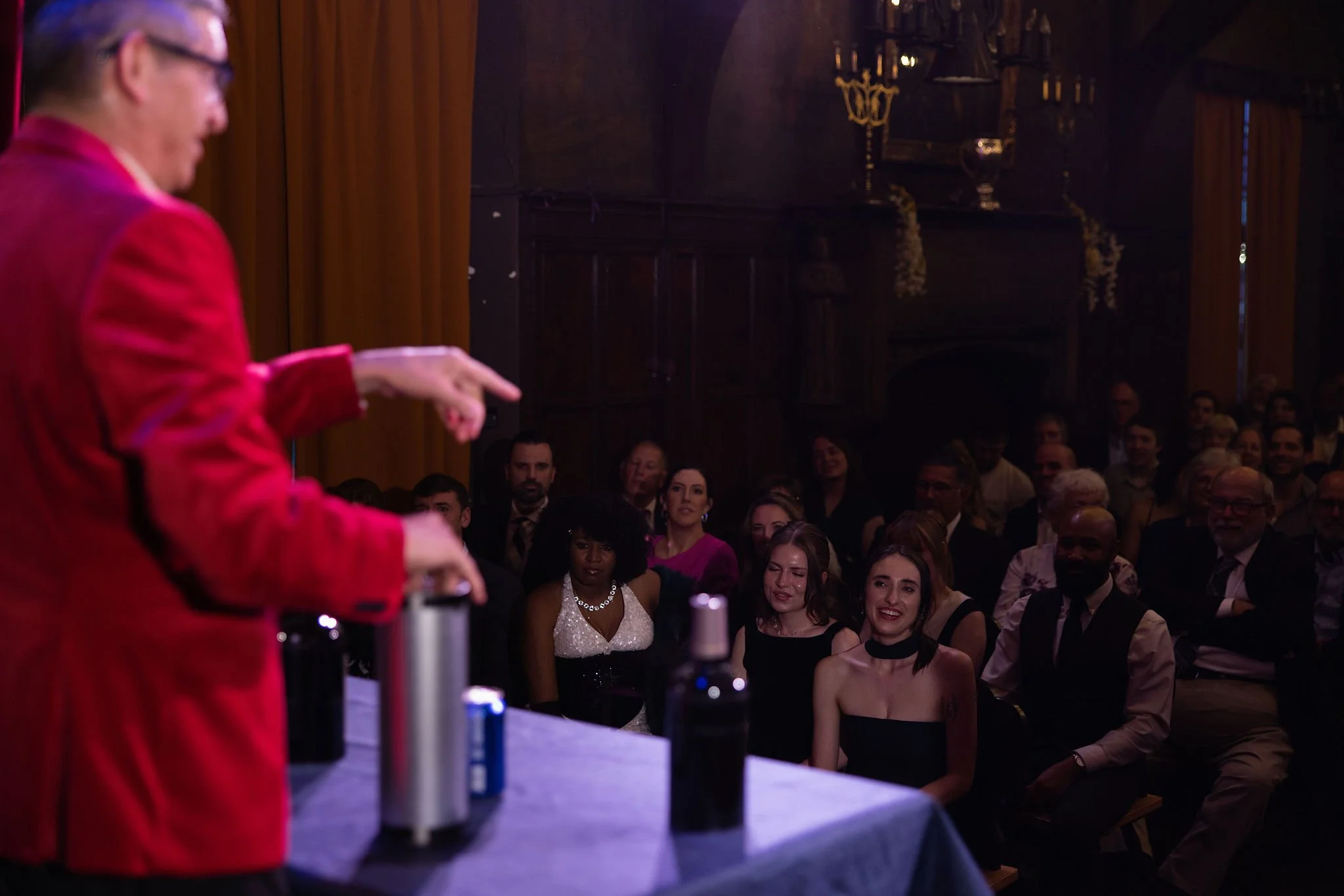 A person in a red jacket points at a table with various bottles in front of an audience in a dimly lit room with wooden paneling and chandeliers.