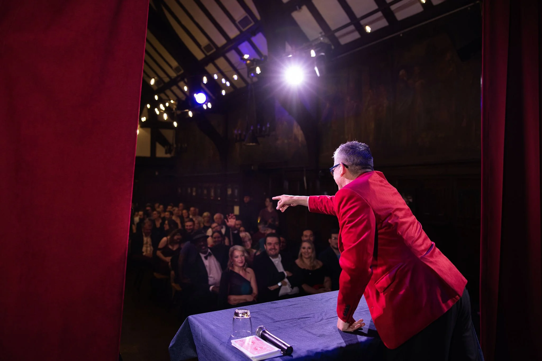 Performer in a red jacket pointing towards the audience during a show in a theater, with an audience seated and smiling in the background.