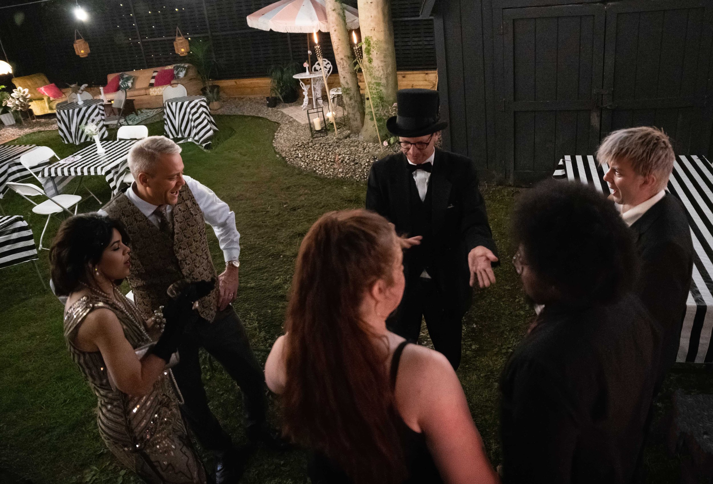 People dressed in formal and vintage costumes celebrating at an outdoor night party, with striped tables and string lights.