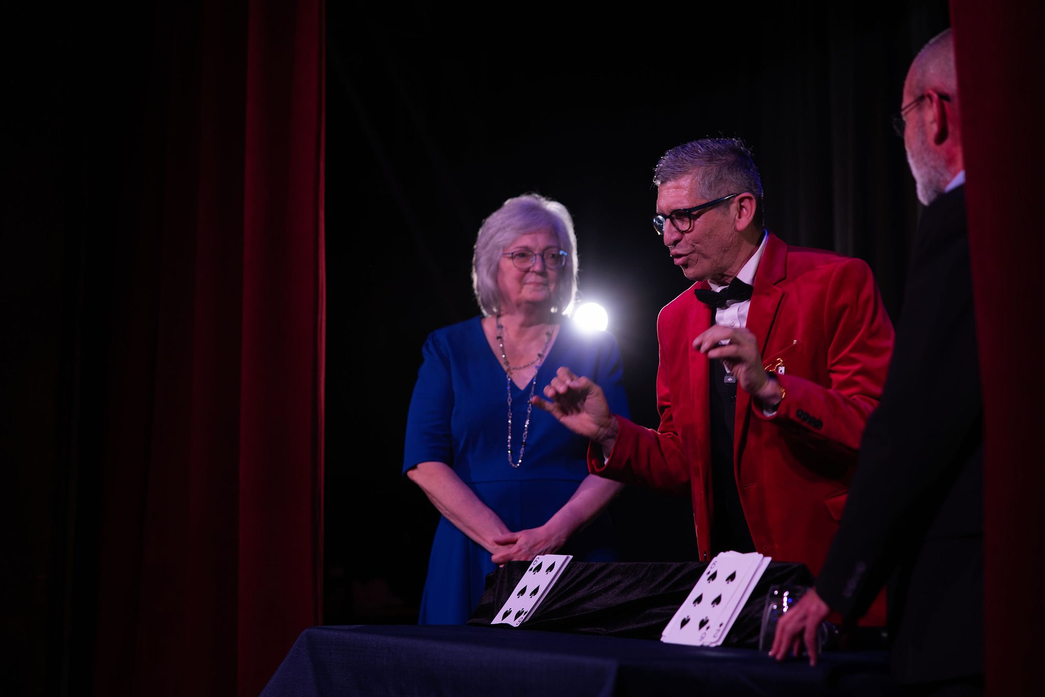 Cleveland magician David Lee in a red jacket performs a card trick with a deck of playing cards on a table, while a woman and other people watch in a dark theater with red curtains.