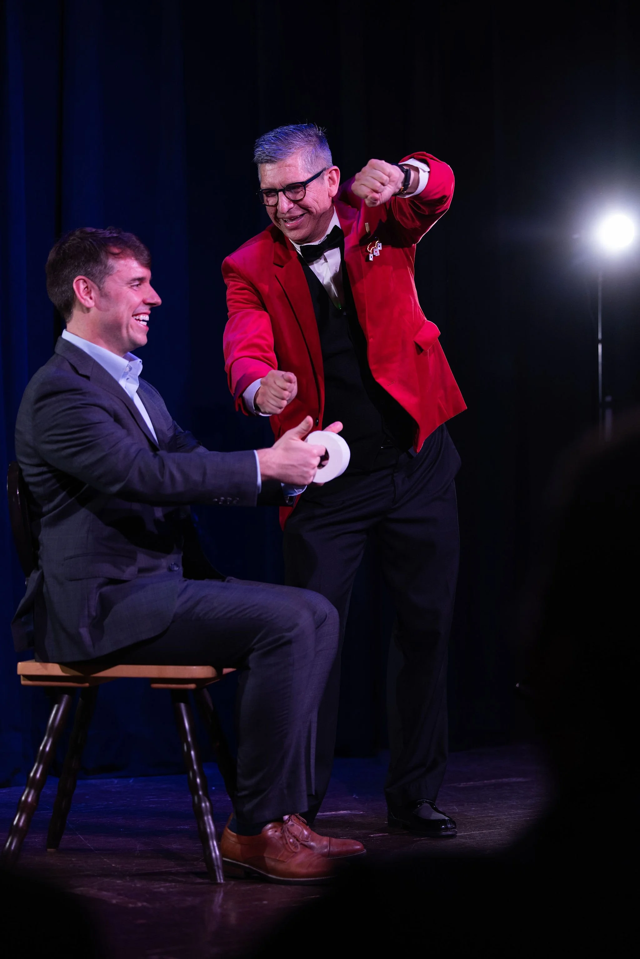 Two men on stage, one sitting on a chair and the other standing, both dressed in formal attire, with stage lighting in the background.