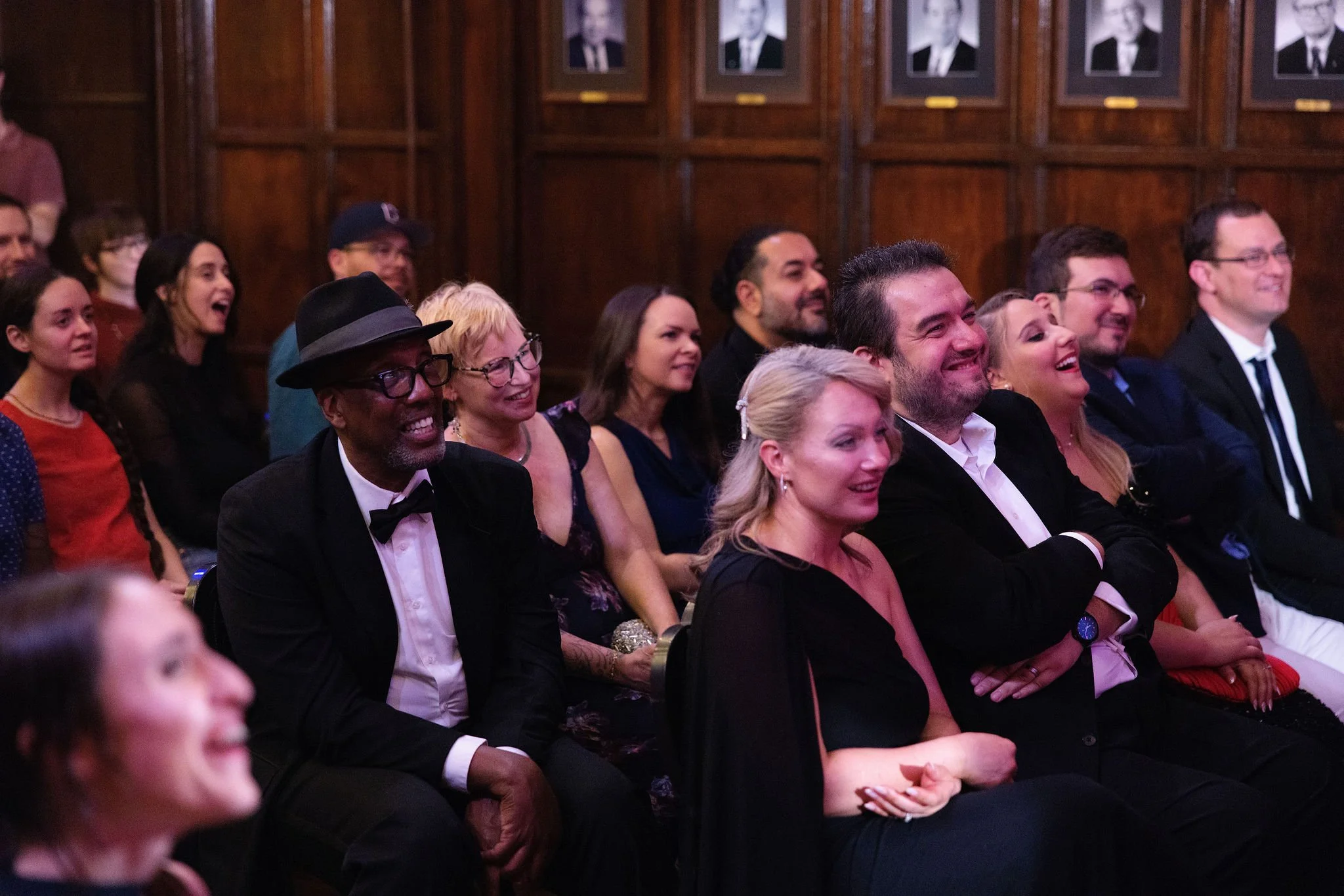 Audience members sitting in seats watching an event, all smiling and laughing in a wood-paneled room with framed black-and-white portraits on the wall.