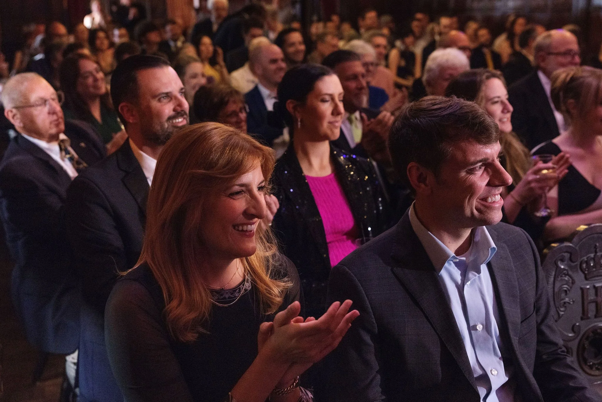 Audience members, including men and women of various ages, seated in rows at a formal event, clapping and smiling.