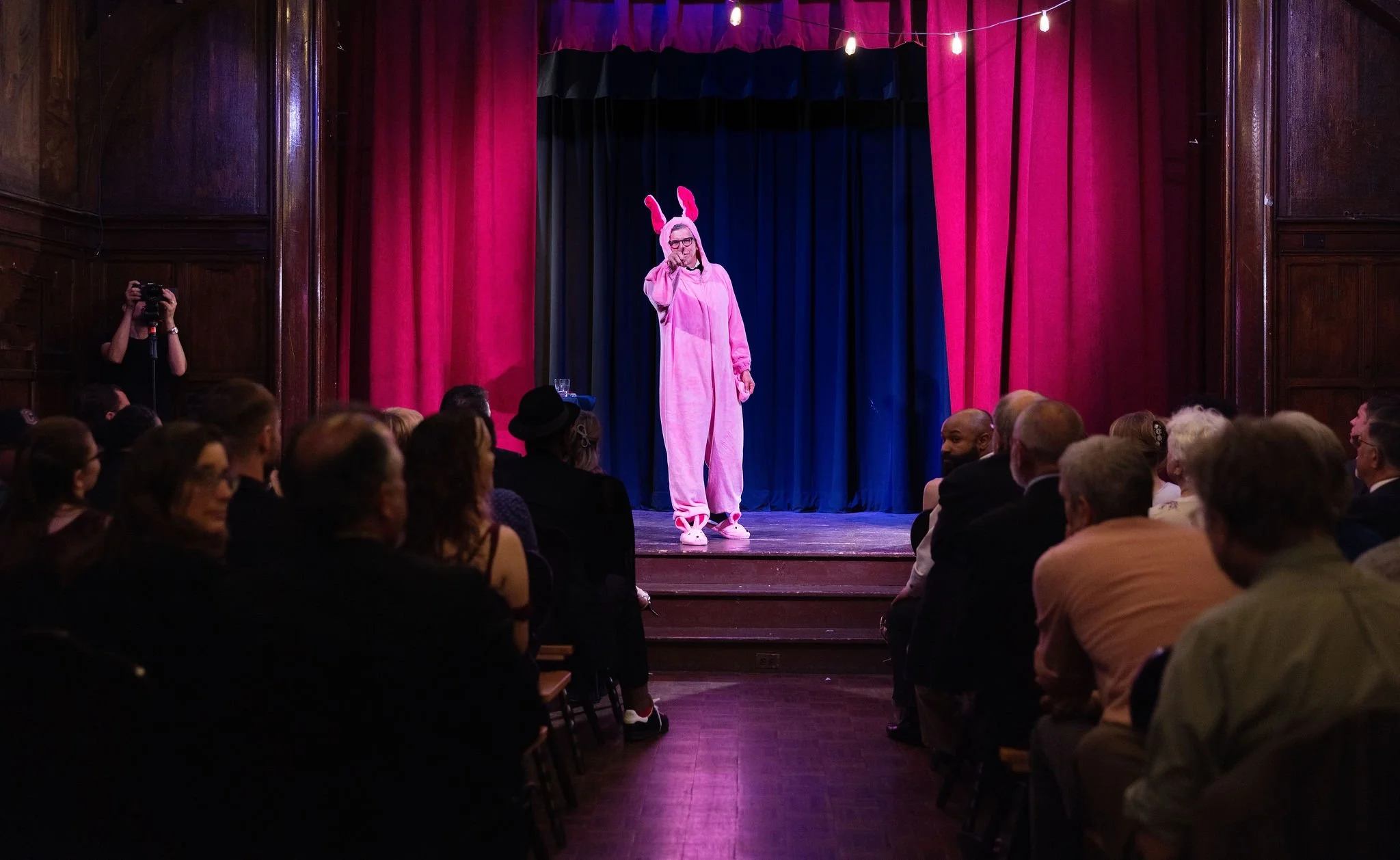 Person dressed in pink bunny costume standing on stage, speaking into microphone, with an audience seated in front and a photographer taking pictures.