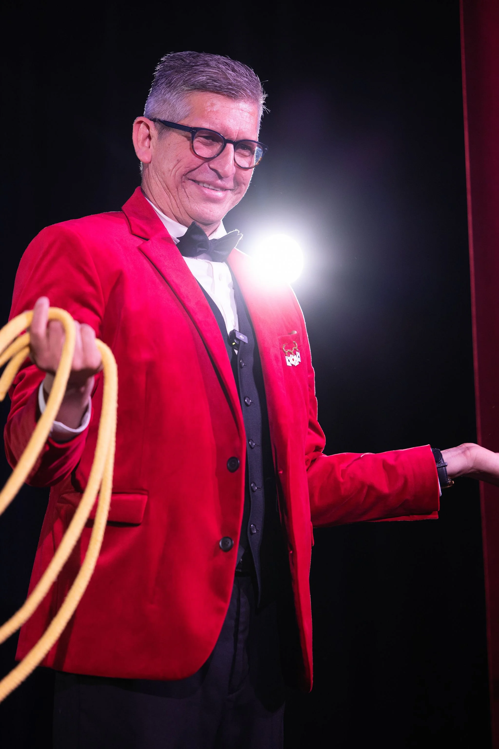 A man dressed in a red tuxedo jacket, white shirt, black bow tie, and glasses, smiling with arms extended, holding a yellow rope in one hand, against a dark background with a bright light glare behind him.