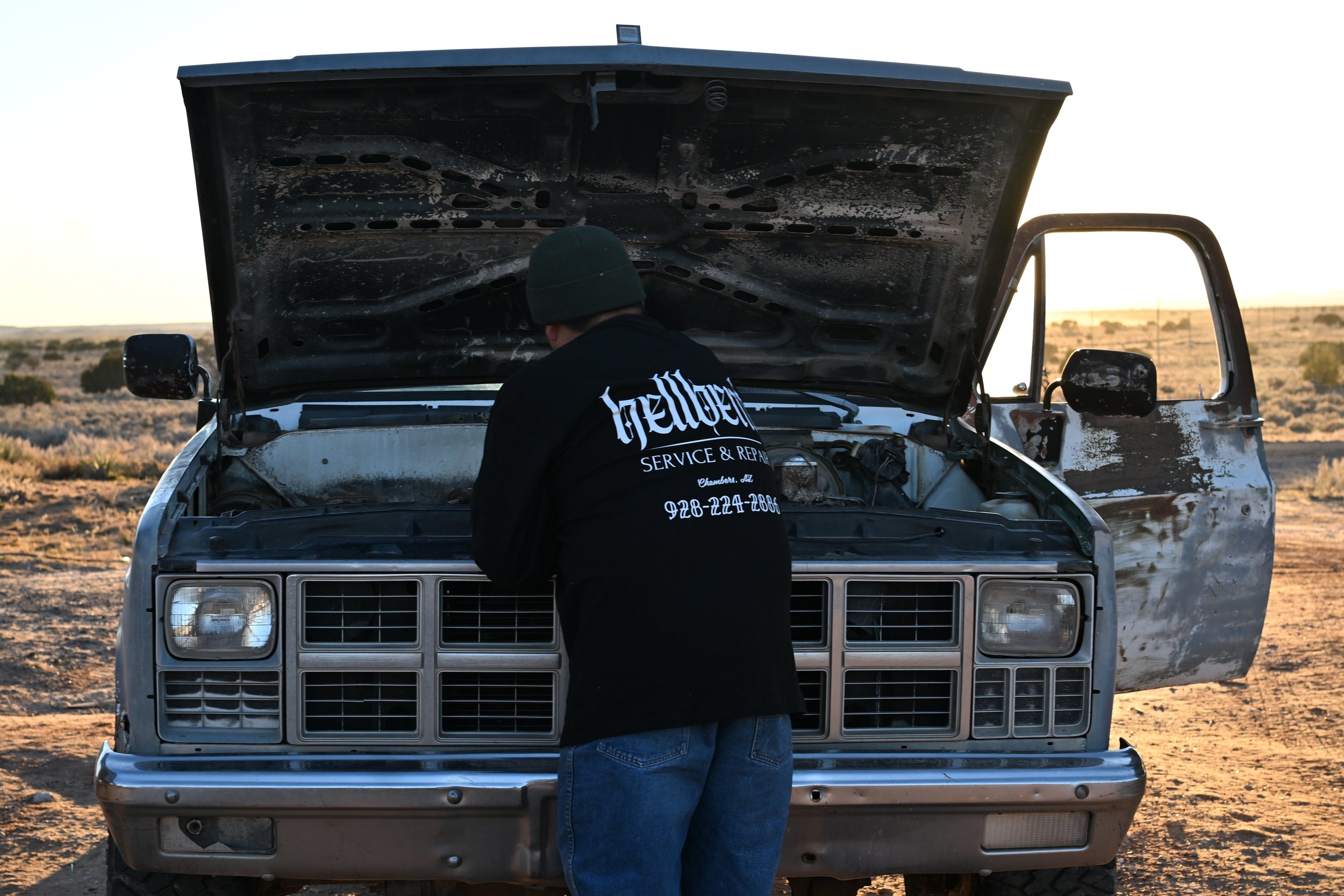 A person wearing a black beanie and a black shirt with Hellbent Service and Repair details on the back stands in front of a vintage white pickup truck with its hood open in a desert landscape during sunset.