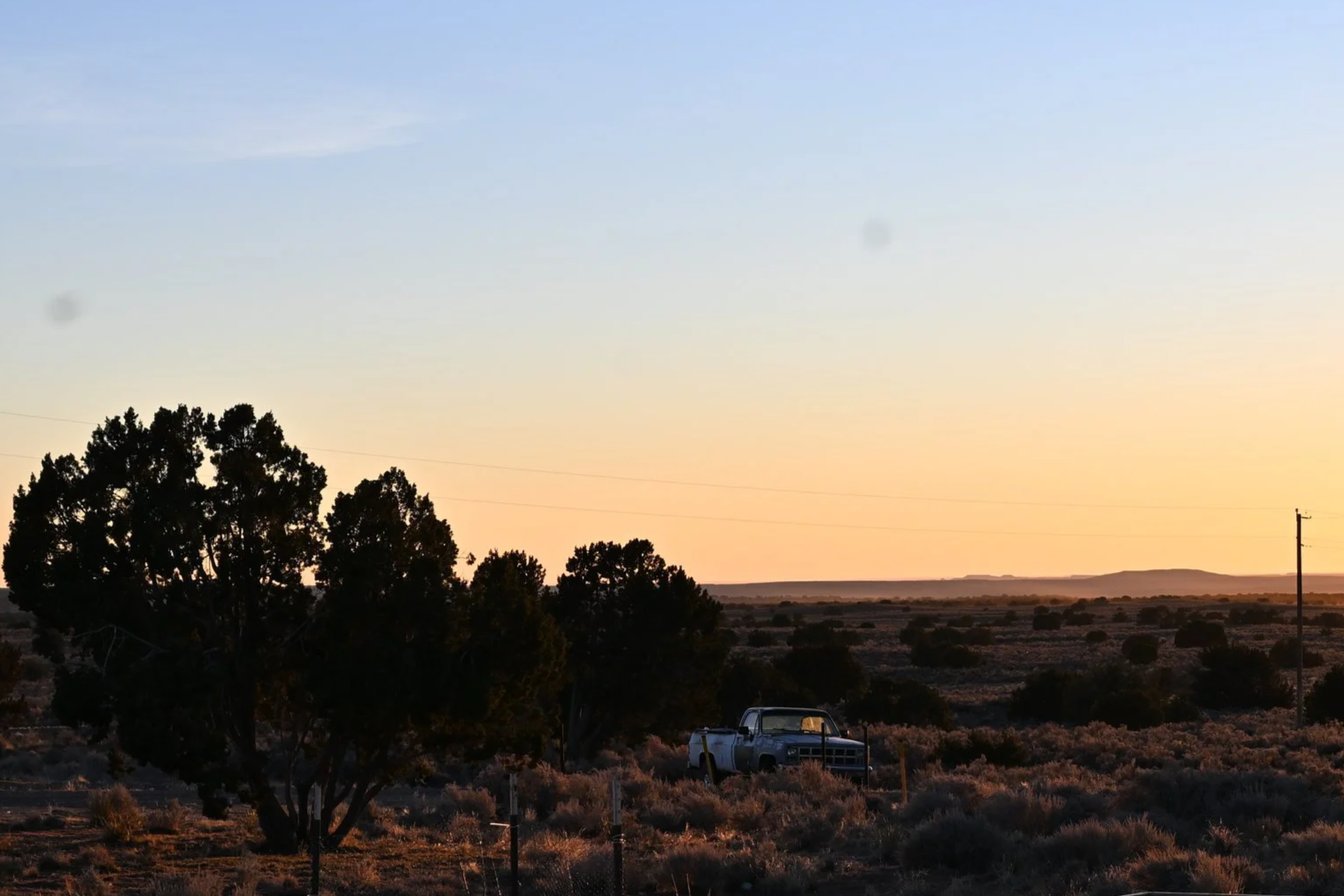 A rural landscape at sunset with a large tree, a white pickup truck, power lines, and distant hills under a clear sky.