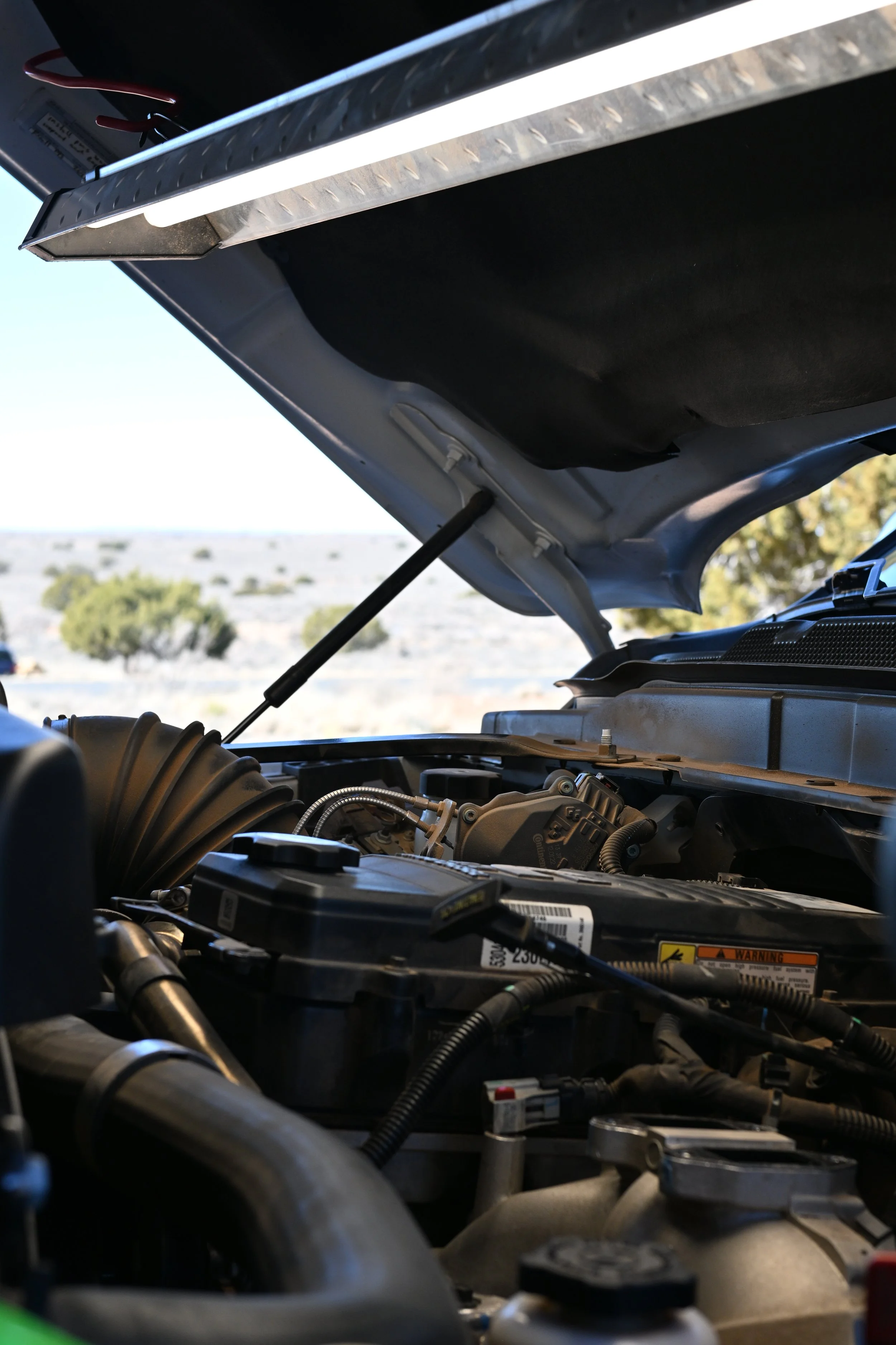 Close-up view of an open car hood revealing engine components, hoses, and wiring against a backdrop of a clear sky and landscape.