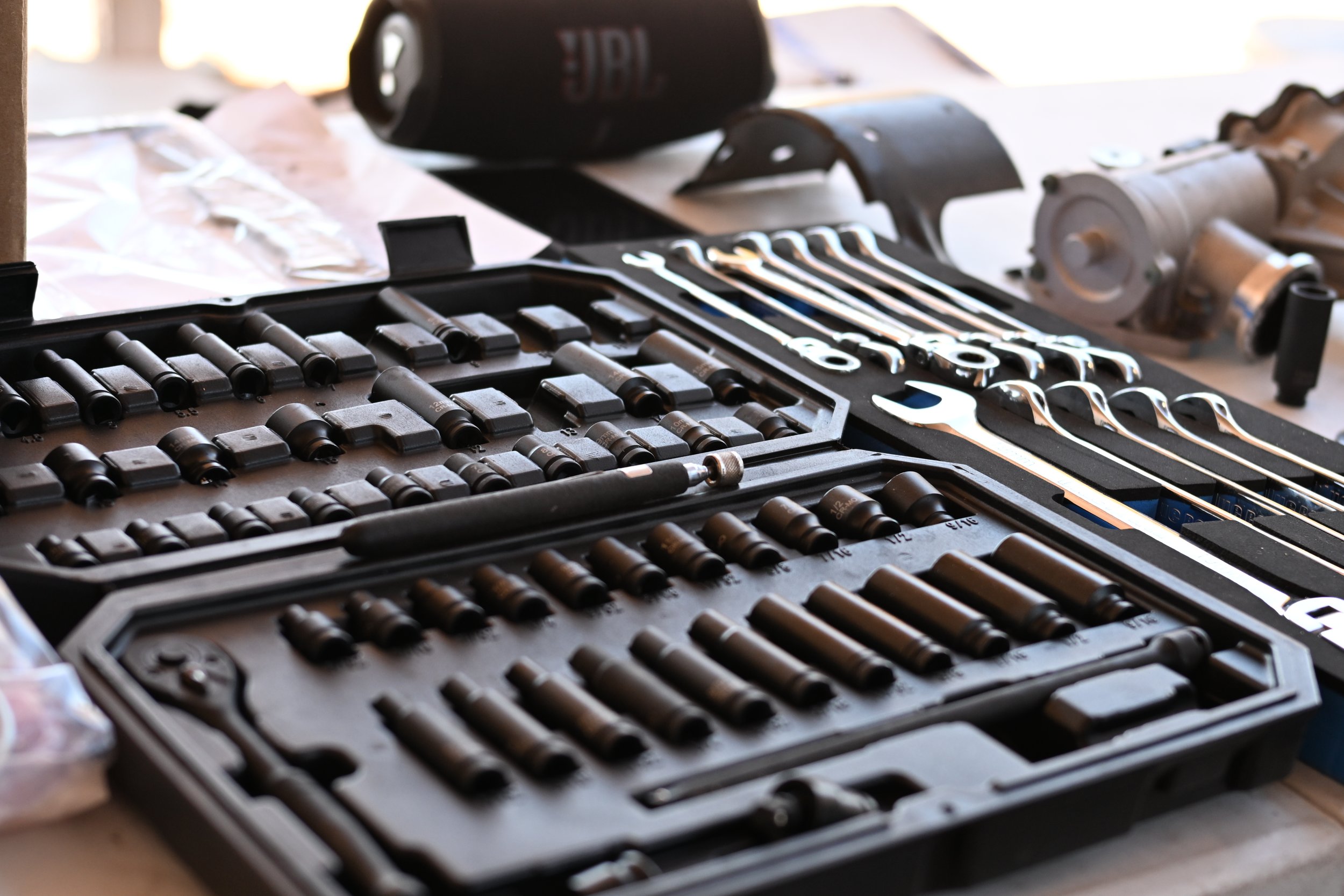 A toolbox containing various sockets, wrenches, and tools laid out on a workbench, with a JBL portable speaker in the background.