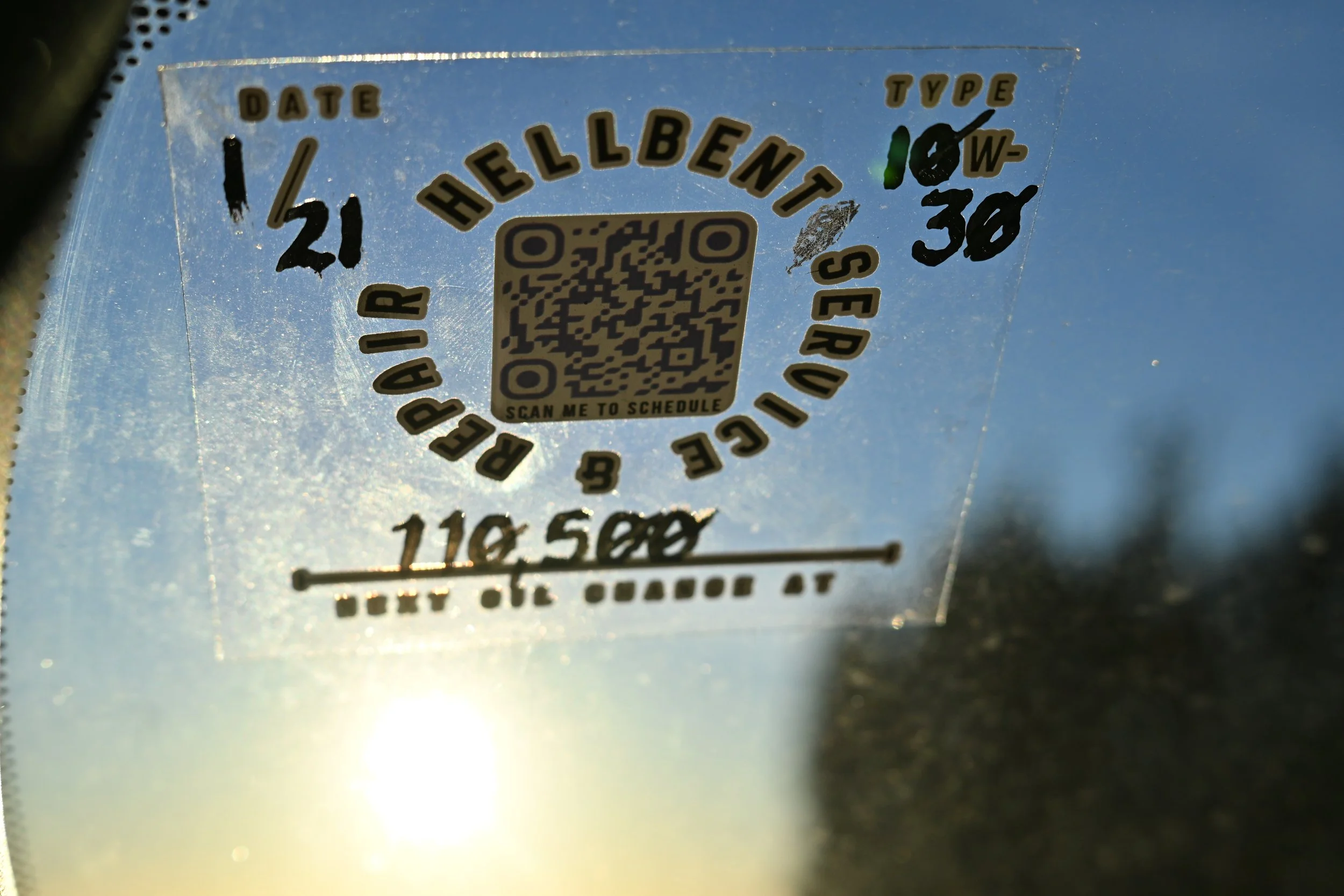 Close-up of a transparent sticker with black and gold text, including a QR code in the center, attached to a reflective surface. The sticker contains various words, dates, and a URL for scheduling.