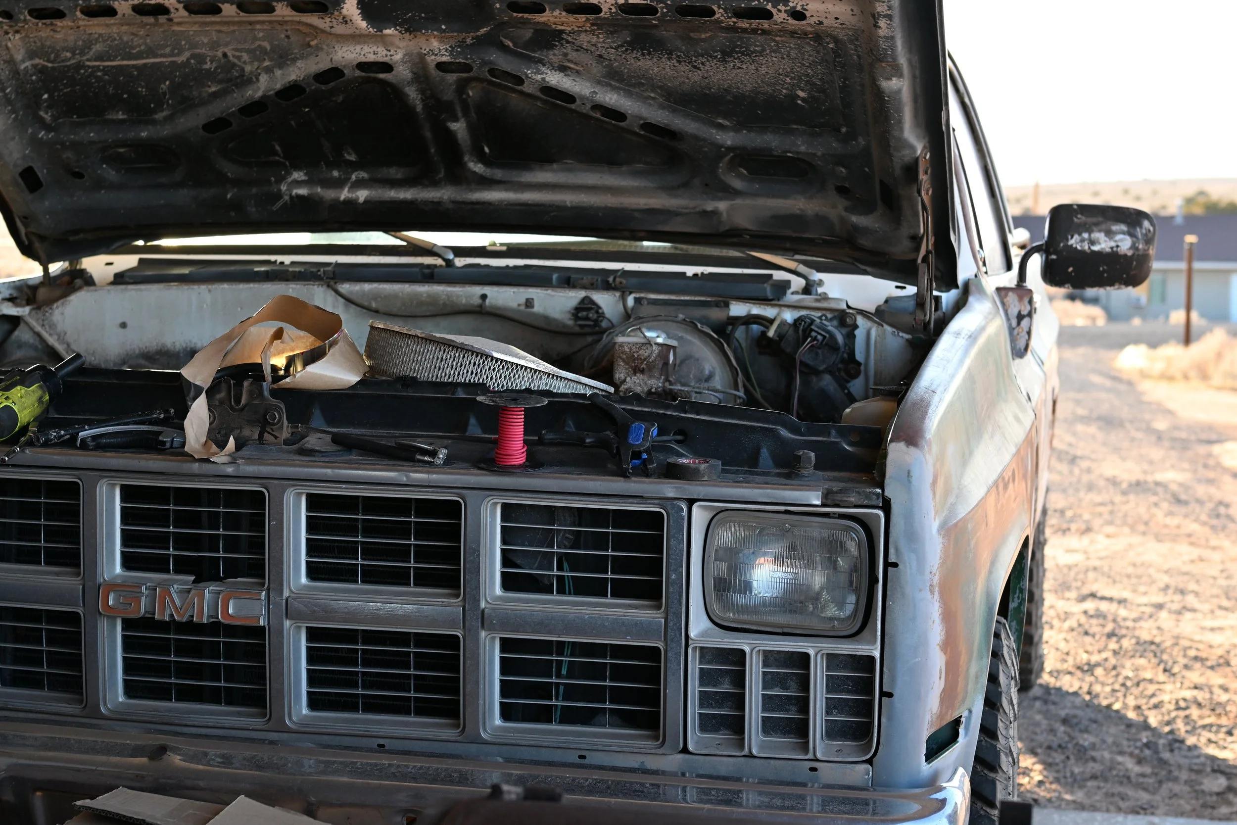 Front view of an old, dusty GMC vehicle with its hood open, exposing the engine bay which contains various parts and tools, in an outdoor setting.