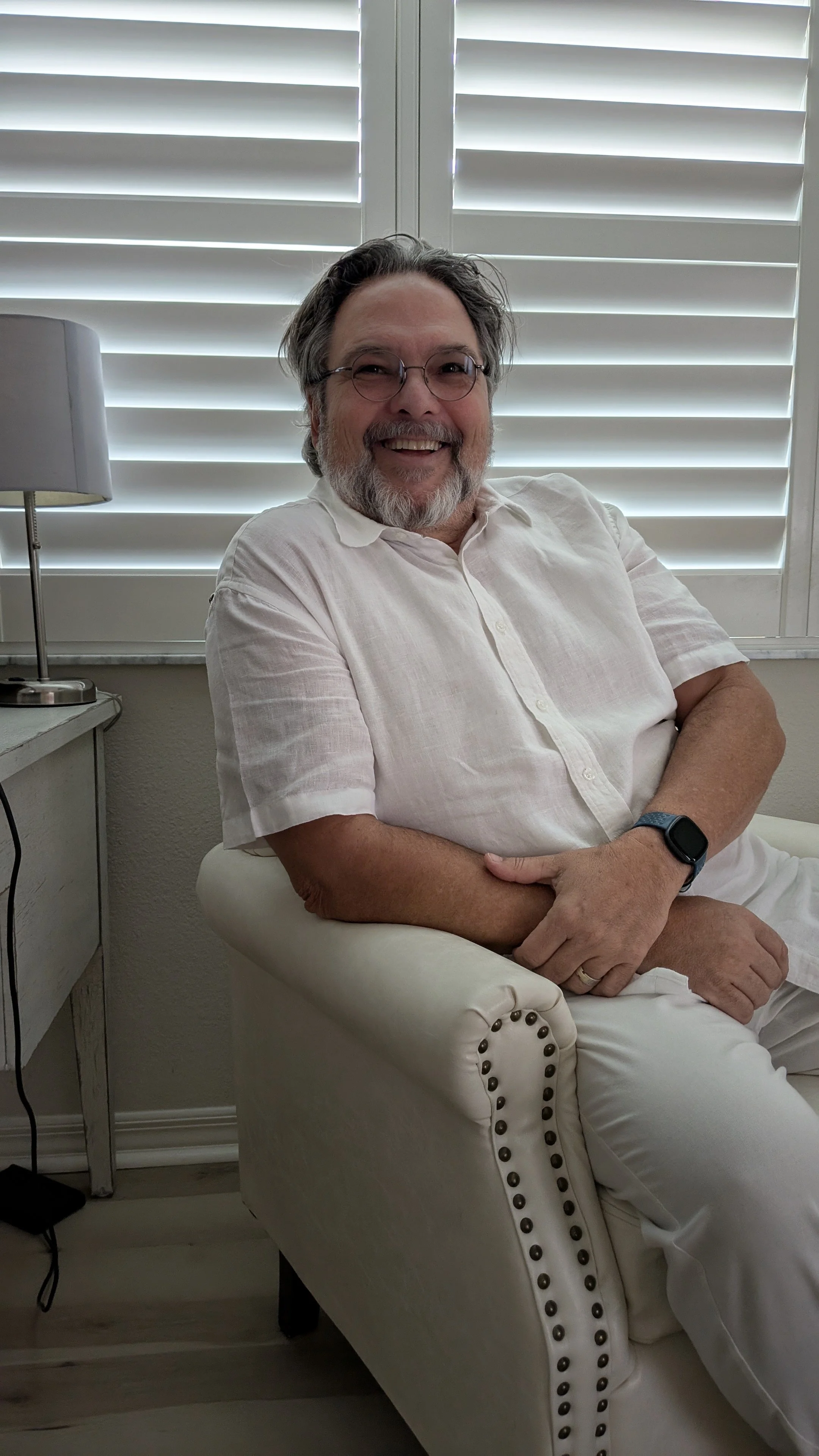 A man with glasses, a beard, and gray hair, wearing a white short-sleeve button-up shirt, sitting on a white armchair in a room with white plantation shutters. He is smiling and has a smartwatch on his left wrist.
