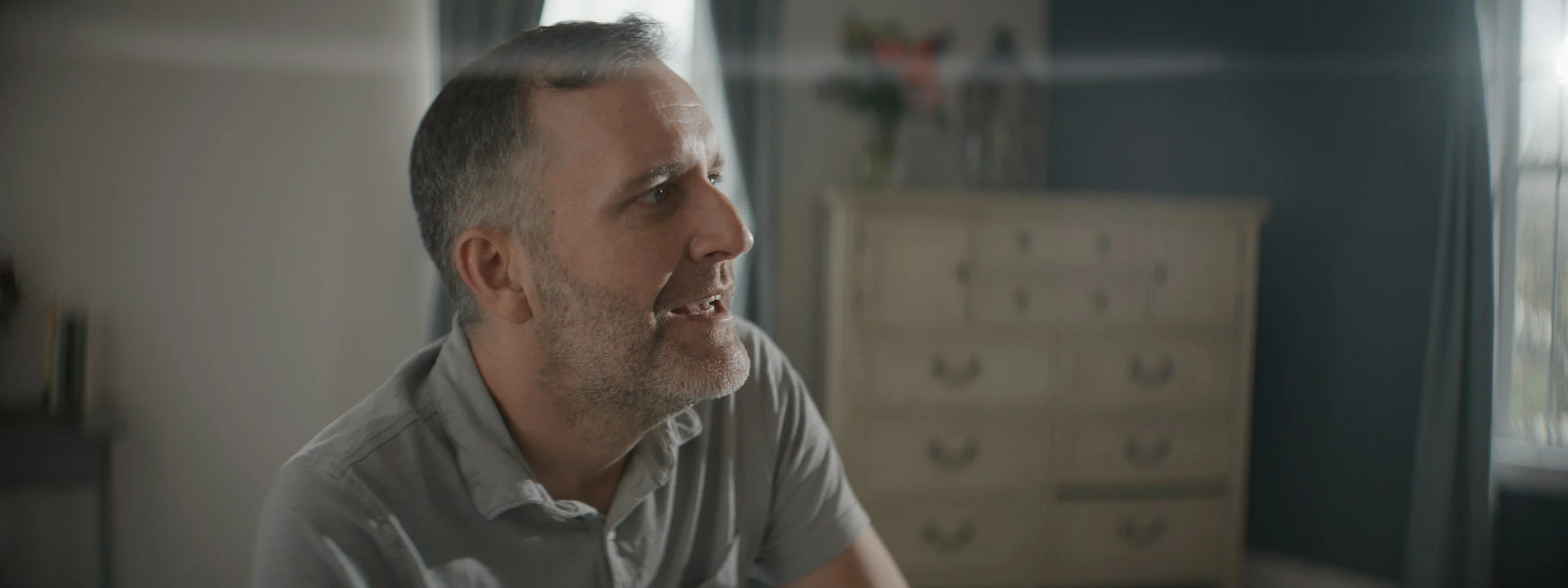 Close-up of a middle-aged man with gray hair and beard, sitting and speaking in a room with natural light and a dresser in the background.
