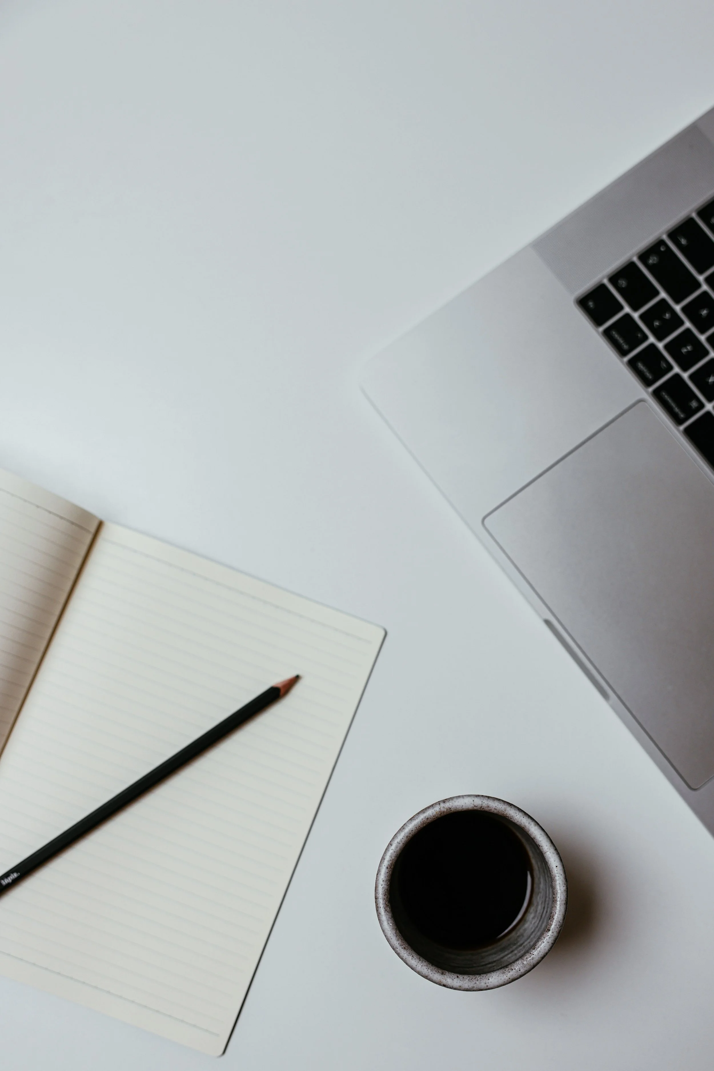 A workspace on a white desk includes an open notebook with lined pages, a black pen, a laptop with a silver body and black keyboard, a cup of black coffee, and a small notepad with a pen resting on top.