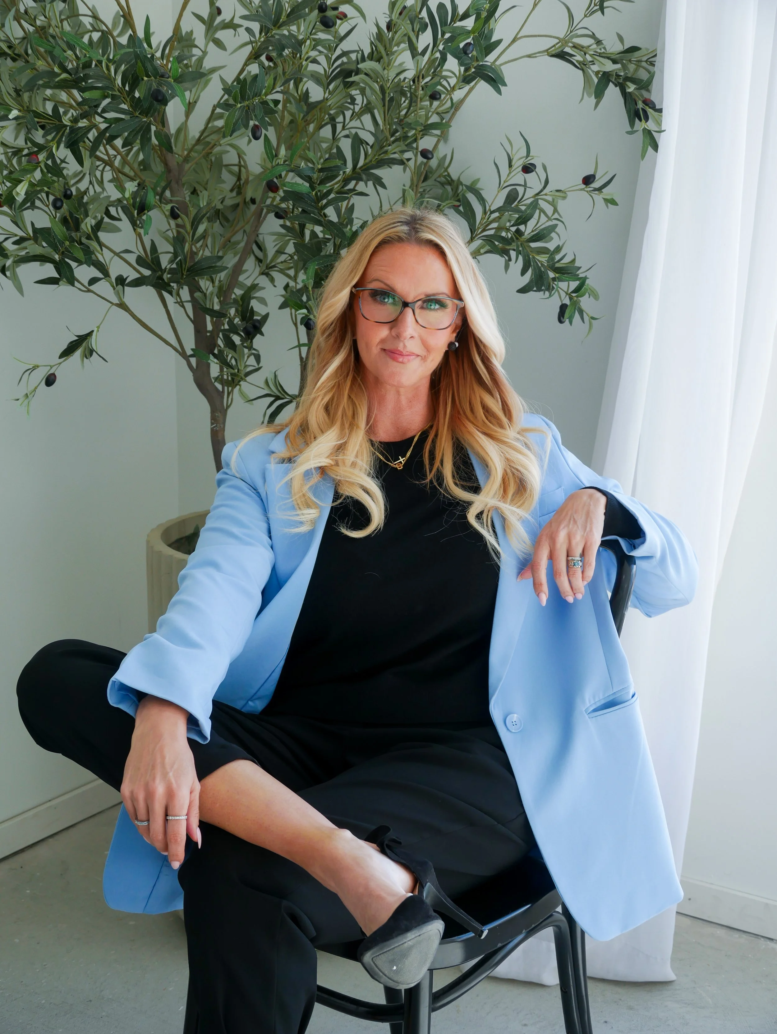 A woman with long blonde hair, glasses, and earrings is sitting on a chair, wearing a black dress and a light blue blazer. She has one leg crossed over her knee and is looking at the camera. Behind her is a leafy bush with dark berries, and white curtains are visible on the right side of the image.
